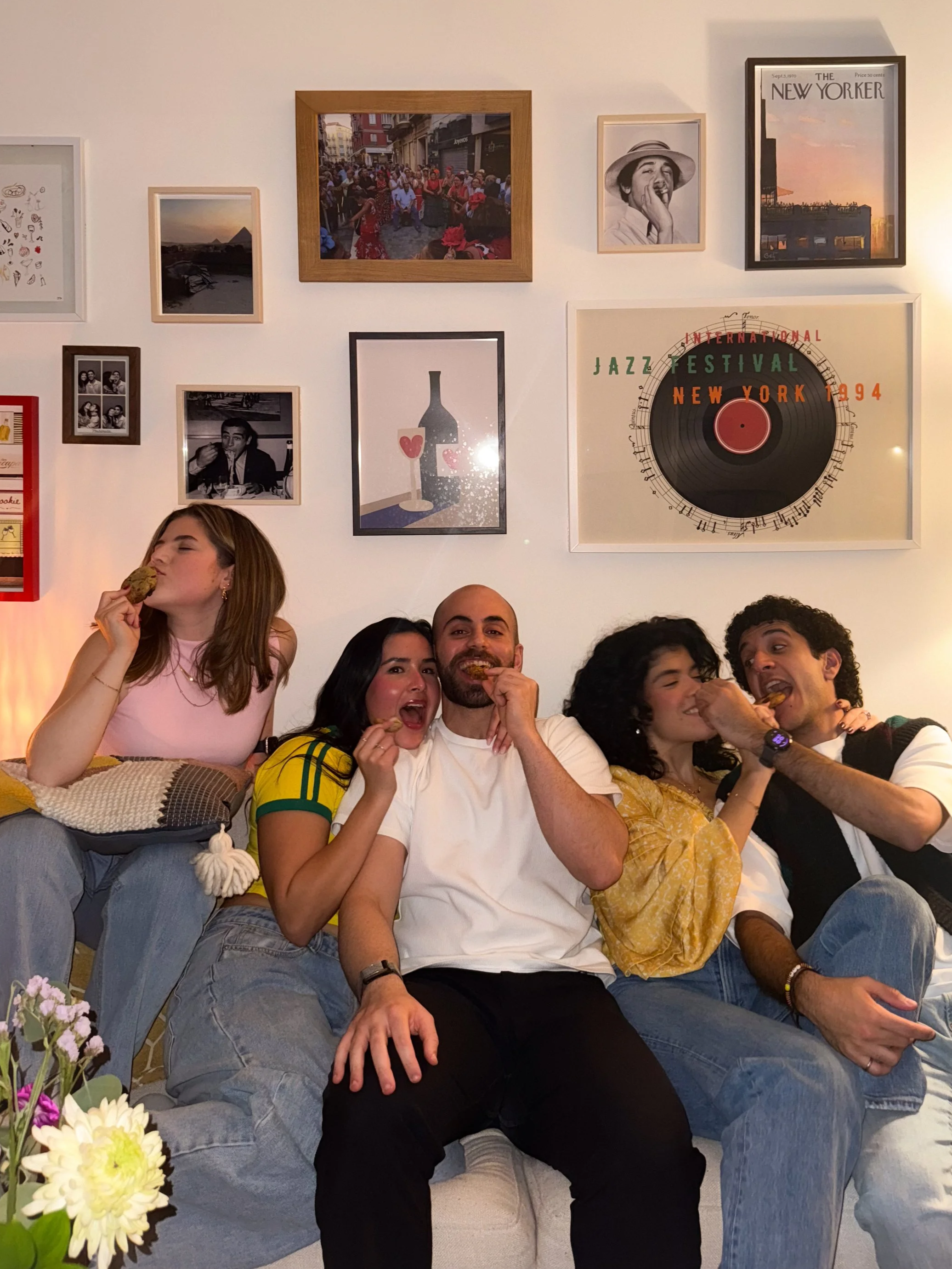 Group of six friends sitting on a couch inside a living room, eating cookies. Behind them, a white wall decorated with framed pictures, including a poster for the New York Jazz Festival 1994. There is a flower arrangement on a table in the foreground.