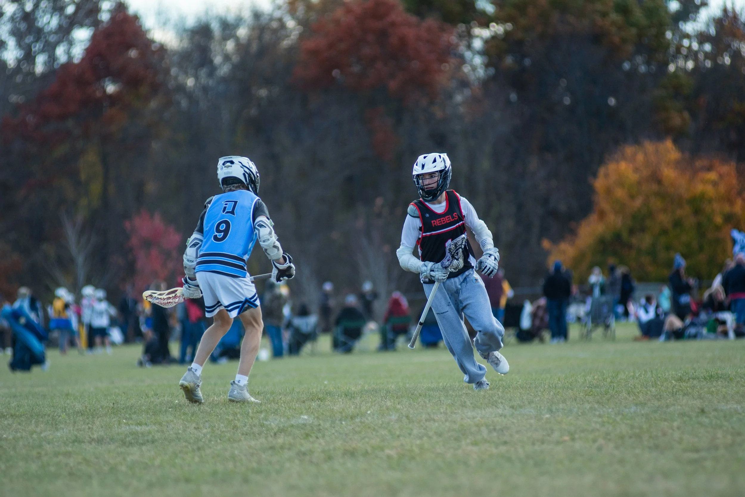 Two young lacrosse players wearing helmets and protective gear playing on a grassy field during fall, with people sitting and standing in the background and colorful autumn trees.