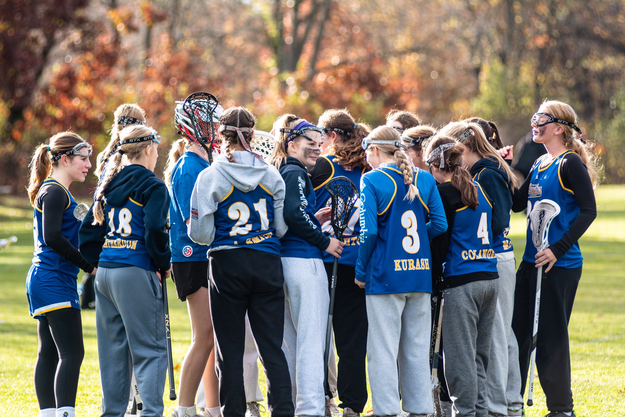 Lacrosse team huddle on a sunny field, wearing blue jerseys and holding lacrosse sticks, with autumn trees in the background.
