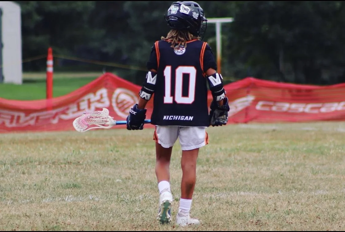 A young lacrosse player wearing a helmet, gloves, and a jersey with the number 10 on the back, standing on a grassy field holding a lacrosse stick, with a red barrier and trees in the background.