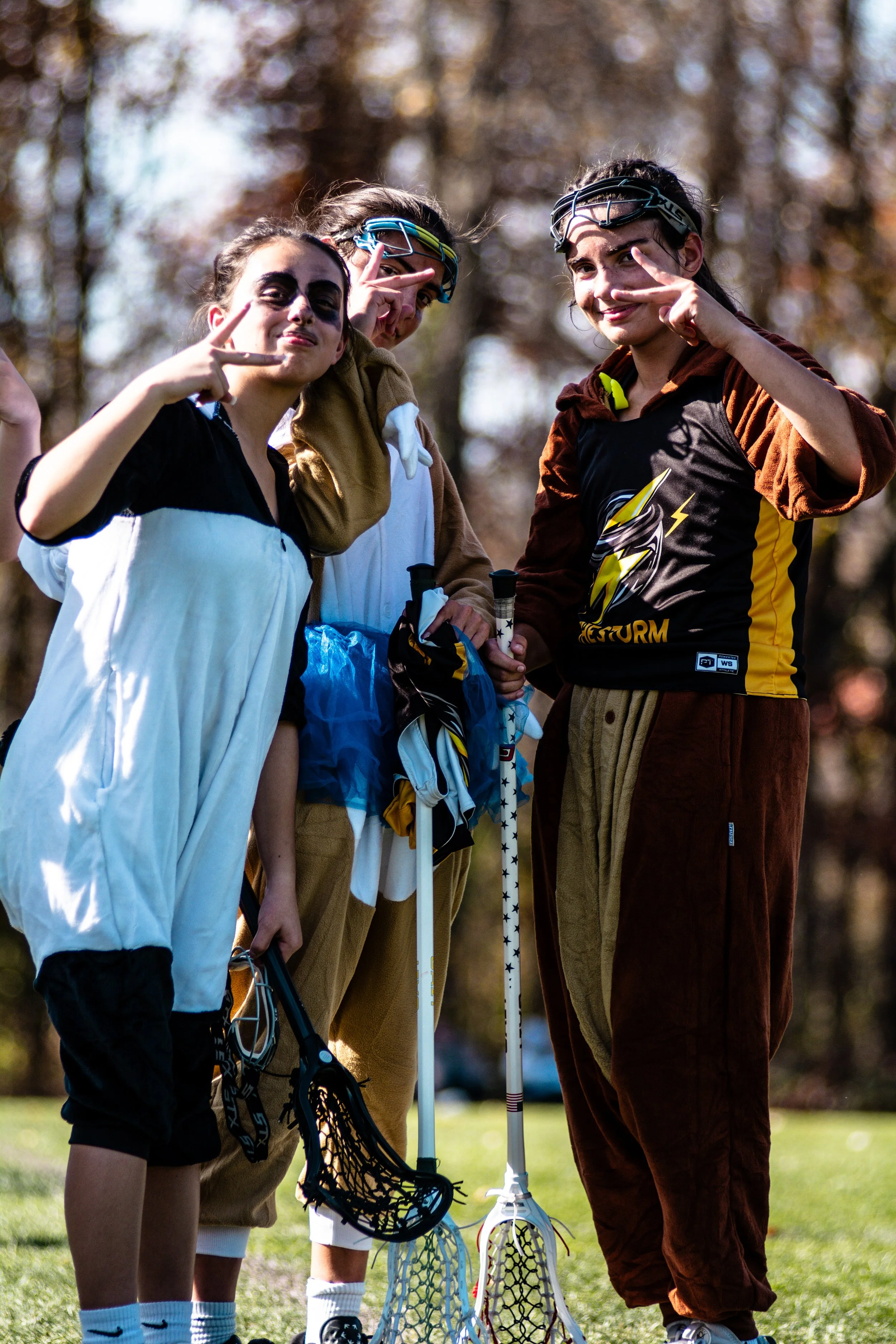 Three young women dressed in costumes with lacrosse sticks, making peace signs and posing outdoors in a park with trees in the background.