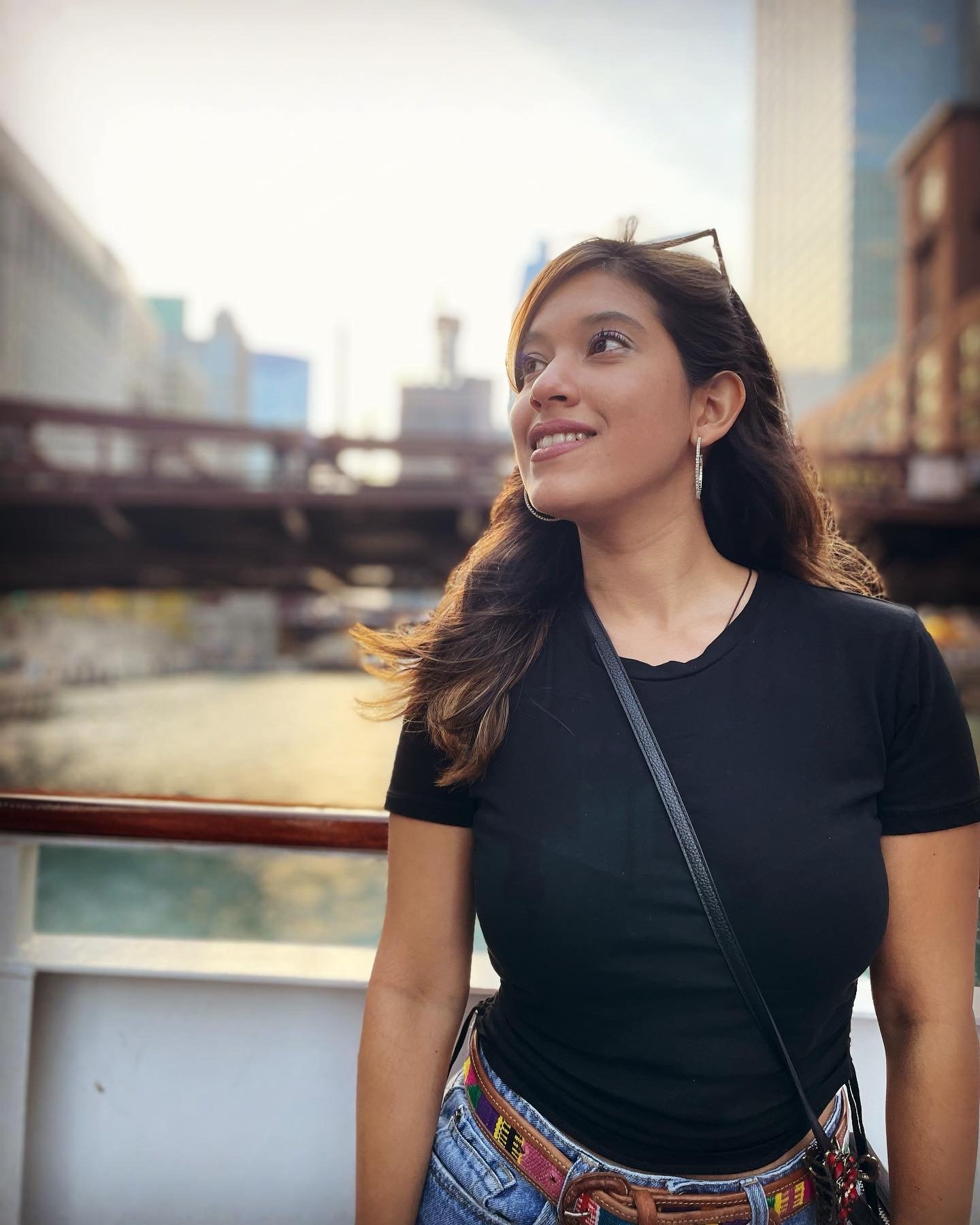 Woman in black shirt standing by a river, with cityscape and bridge in the background.