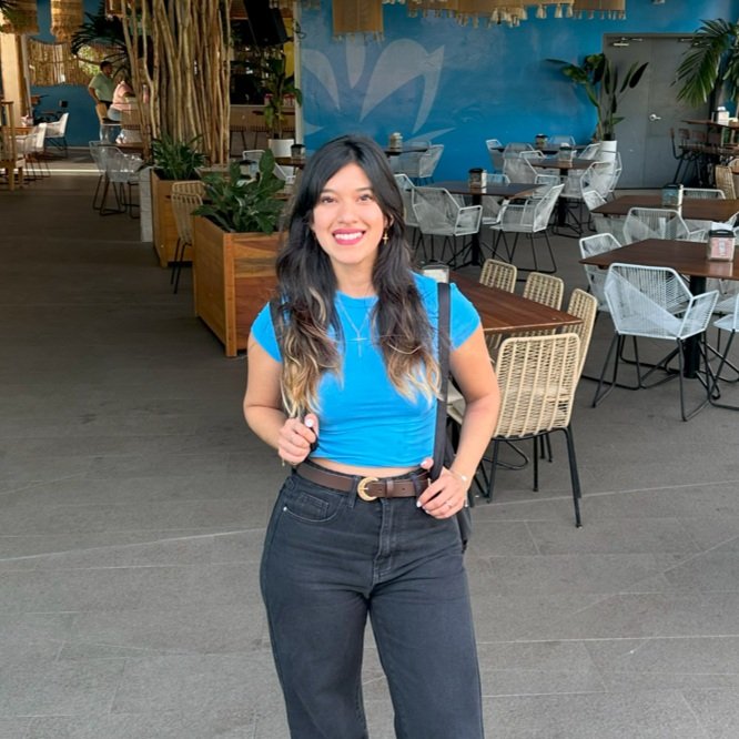 Woman in a blue shirt and black pants, standing in a restaurant with empty chairs and tables in the background.