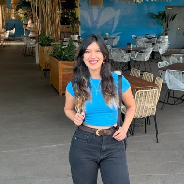 Woman in a blue shirt and black pants smiling in a restaurant setting with tables, chairs, and plants in the background.