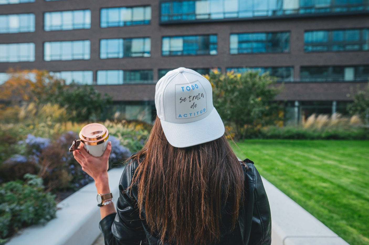 Woman wearing a leather jacket and a white cap with Spanish text, holding a travel mug, standing outdoors in front of a modern building with green grass and plants.