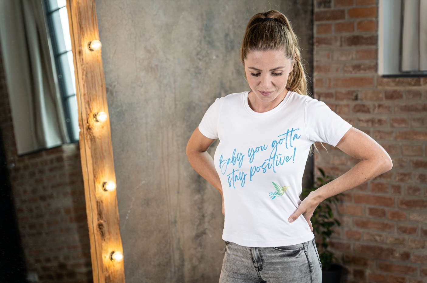 Person wearing a white t-shirt with "Baby you gotta stay positive!" text standing in front of a mirror with exposed brick and light bulbs.