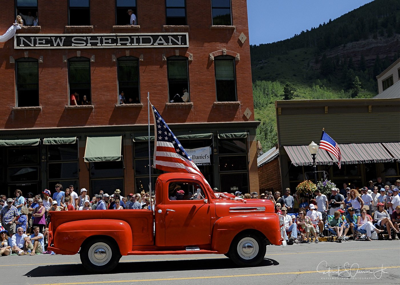A bright red vintage pickup truck with an American flag on top, driving in front of a crowd of spectators during a parade. The background features a historic brick building labeled "New Sheridan" and a mountainous landscape.