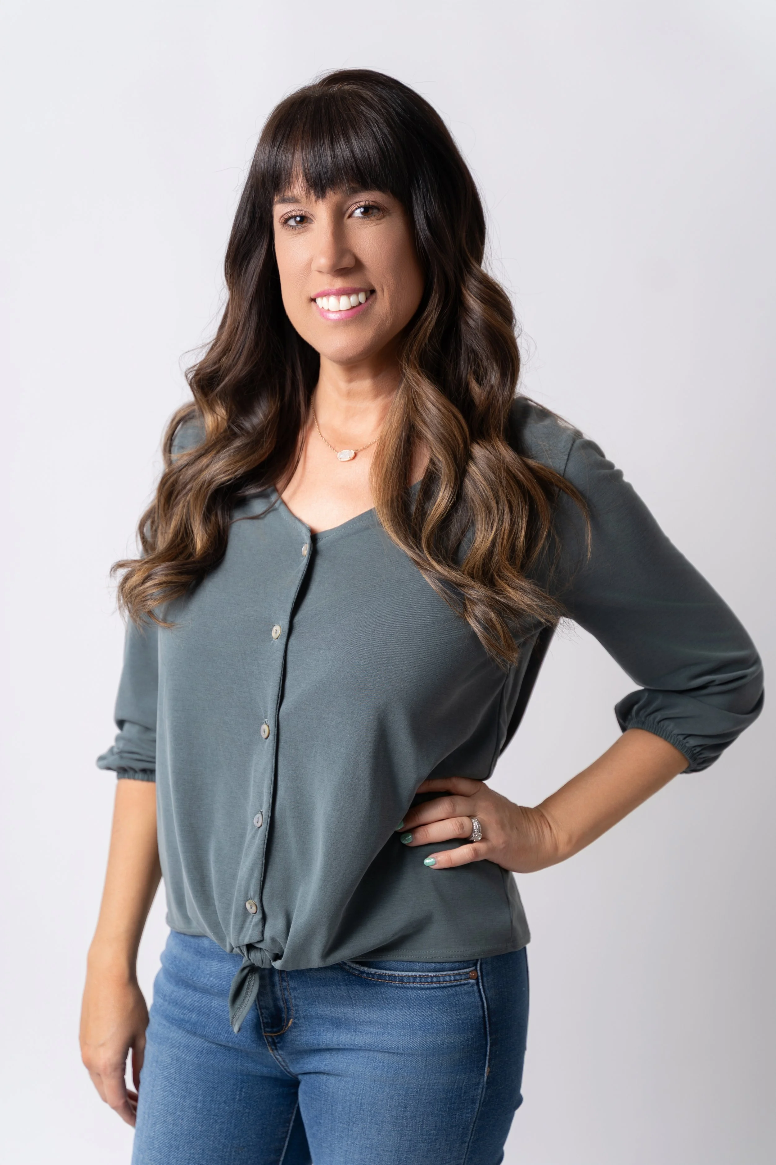 A woman with long, wavy brown hair and a fringe, smiling and posing with one hand on her hip, wearing a gray button-up shirt and blue jeans against a plain white background.