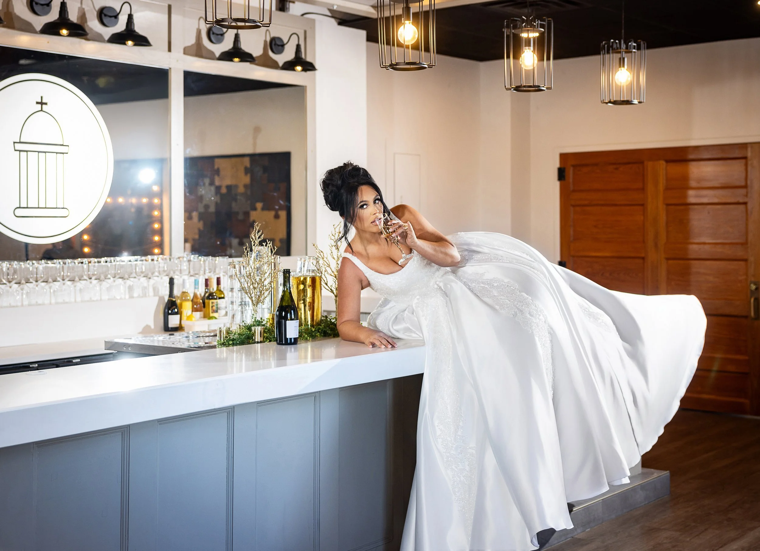 A woman in a white wedding dress playfully lying on a bar counter, sipping a glass of champagne. The bar features several bottles, decorative plants, and hanging light fixtures. Wedding Venue: The Loretto Events -- an upscale event venue and chapel