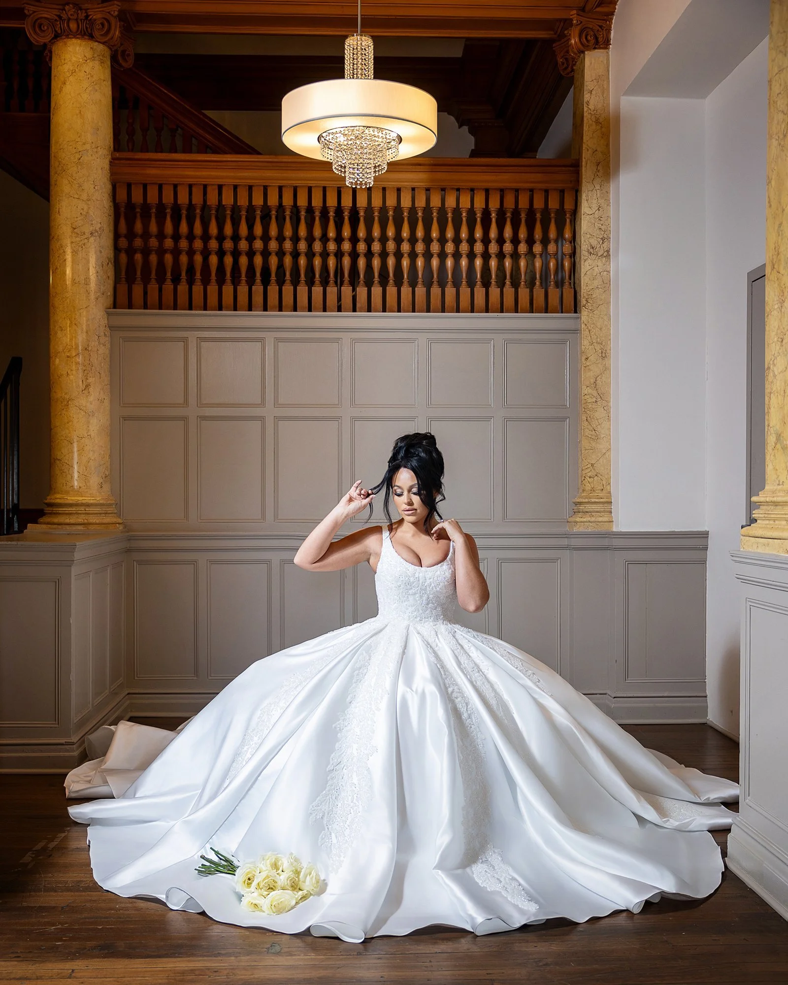 A bride in a luxurious white wedding gown poses indoors with a bouquet of yellow roses on the floor. The setting features elegant wood paneling, columns, and a chandelier. Wedding Venue: The Loretto Events -- an upscale event venue and chapel locate