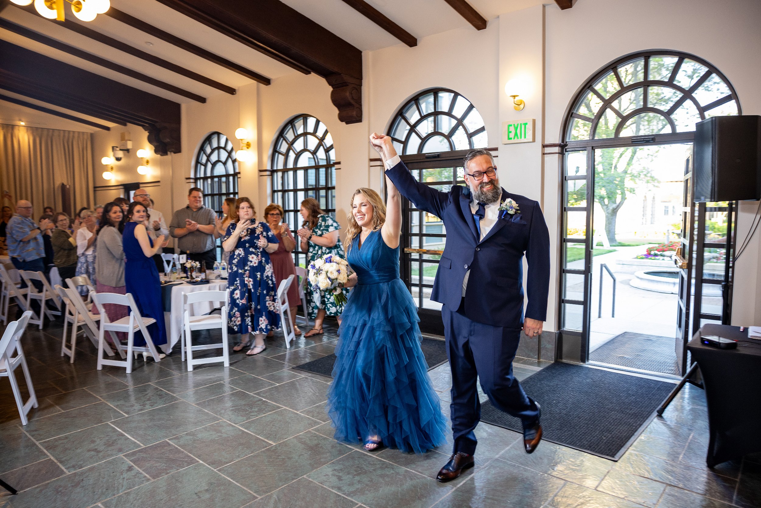 A couple dressed in formal attire enters a reception hall, holding hands joyfully. Wedding Venue: Unity Village -- Experience old European elegance and natural beauty -- Located in Lees Summit, MO.
