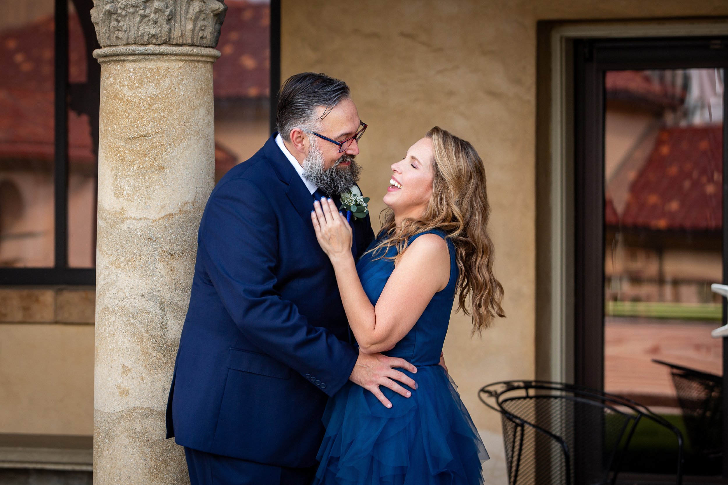 A couple dressed in formal attire smiling and embracing near an ornate column. Wedding Venue: Unity Village -- Experience old European elegance and natural beauty -- Located in Lees Summit, MO.