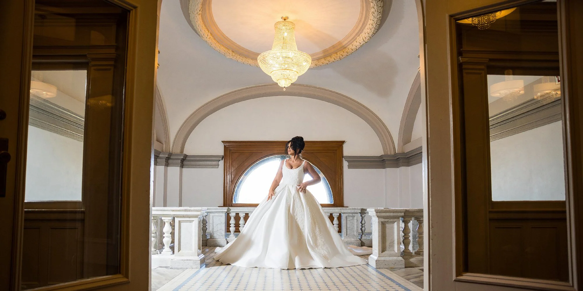 Bride in an elegant white wedding dress standing in a grand hallway with an ornate chandelier and arched windows, viewed through an open doorway. Wedding Venue: The Loretto Events -- an upscale event venue and chapel located in Kansas City, Missouri