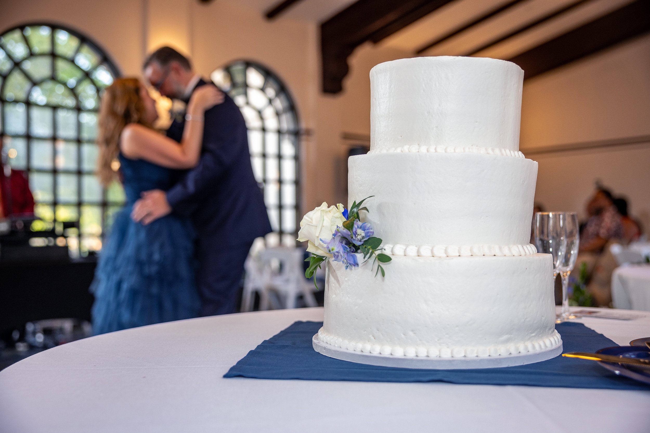 Wedding cake with a floral decoration in the foreground, blurred couple embracing in the background, arched windows. Wedding Venue: Unity Village -- Experience old European elegance and natural beauty -- Located in Lees Summit, MO.