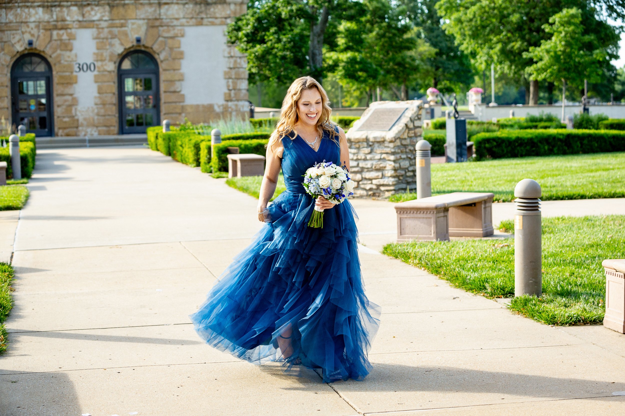 Woman in a blue gown holding a bouquet of flowers, walking outdoors on a pathway, with a historic building and greenery in the background. Wedding Venue: Unity Village -- Experience old European elegance and natural beauty -- Located in Lees Summit,
