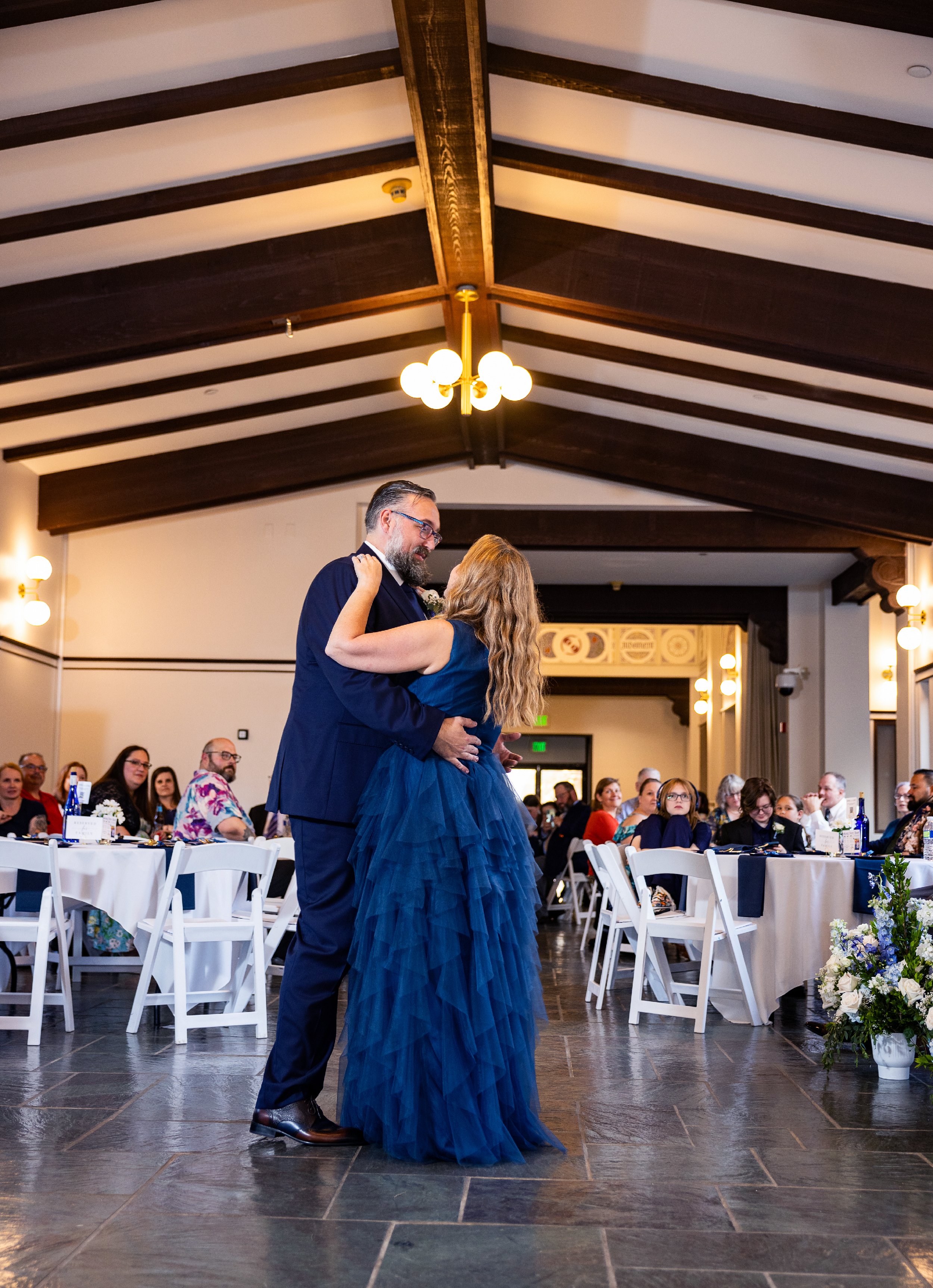 A couple dancing together at a wedding reception in a venue with exposed wooden beams and chandeliers. Guests are seated at tables, watching and enjoying the event. Wedding Venue: Unity Village -- Located in Lees Summit, MO