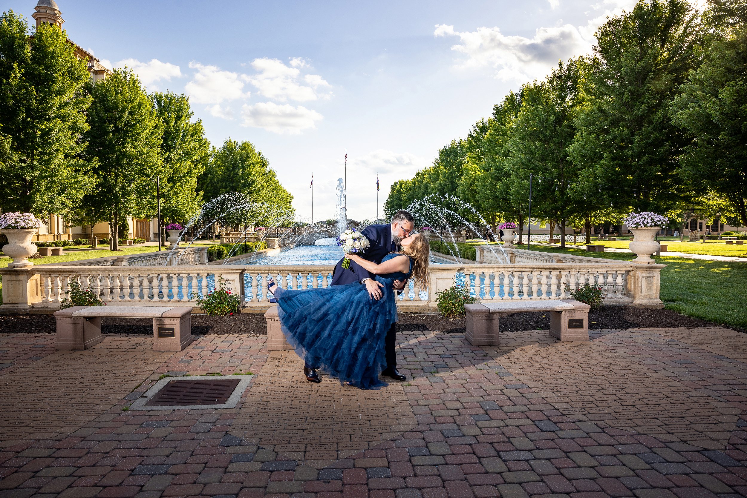 Groom dips bride near a decorative fountain with trees and benches in the background. Wedding Venue: Unity Village -- Experience old European elegance and natural beauty -- Located in Lees Summit, MO.