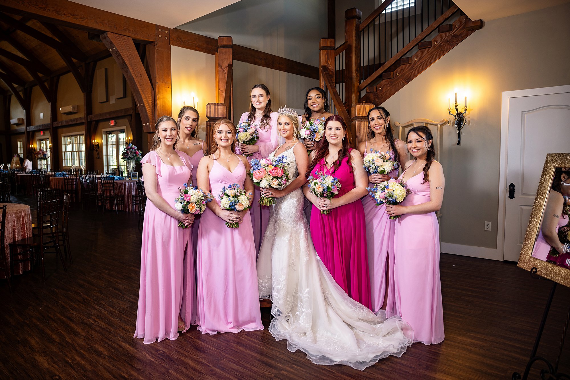 Bride with bridesmaids in pink dresses holding bouquets, standing in a decorated venue. Wedding Venue: The English Barn, a one-of-a-kind castle-inspired venue full of rustic elegance and charm located in Louisburg, KS -- just South of KC.