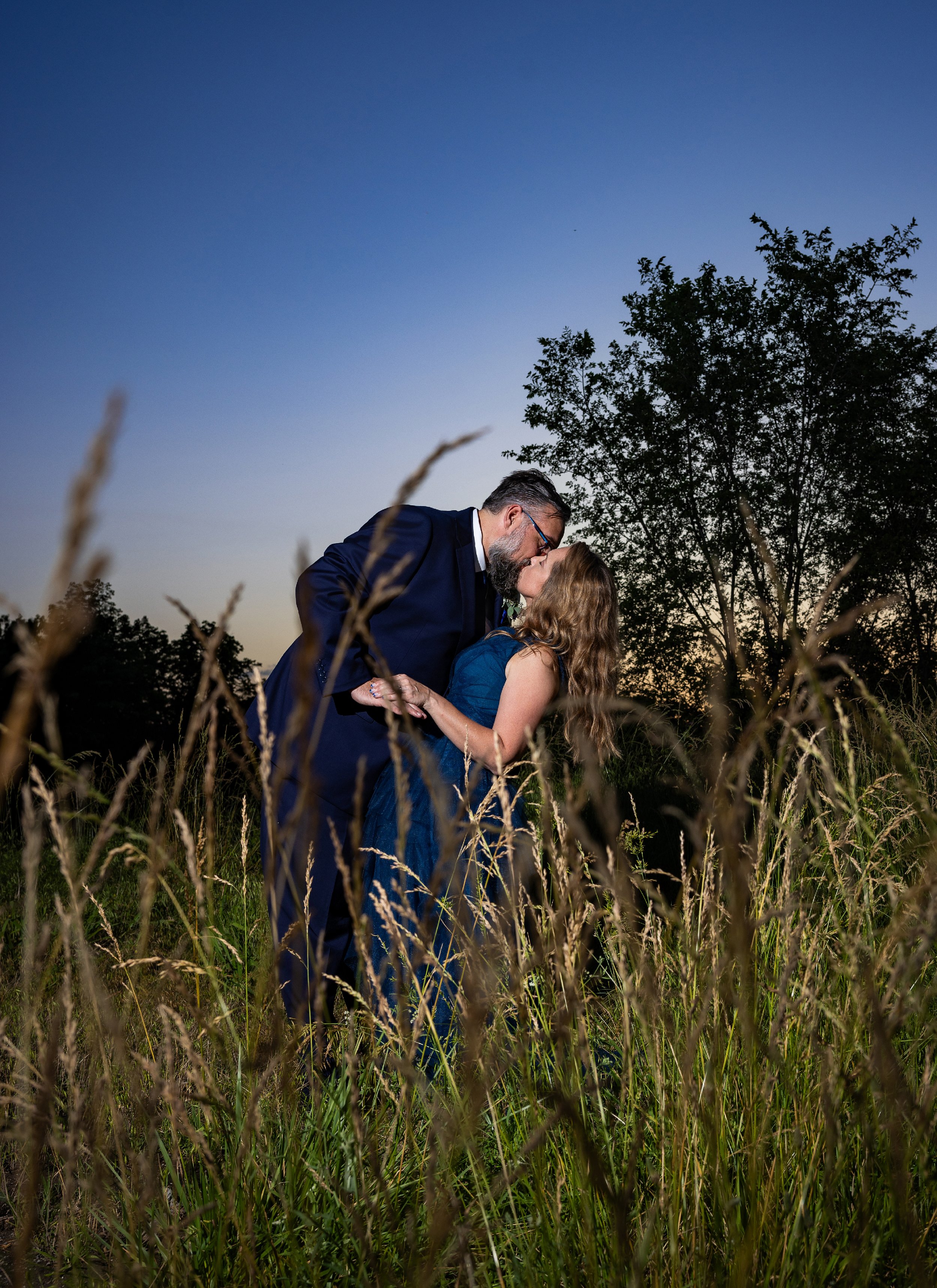 Couple kissing in a field during sunset, surrounded by tall grass, with a tree and blue sky in the background. Wedding Venue: Unity Village -- Experience old European elegance and natural beauty -- Located in Lees Summit, MO.
