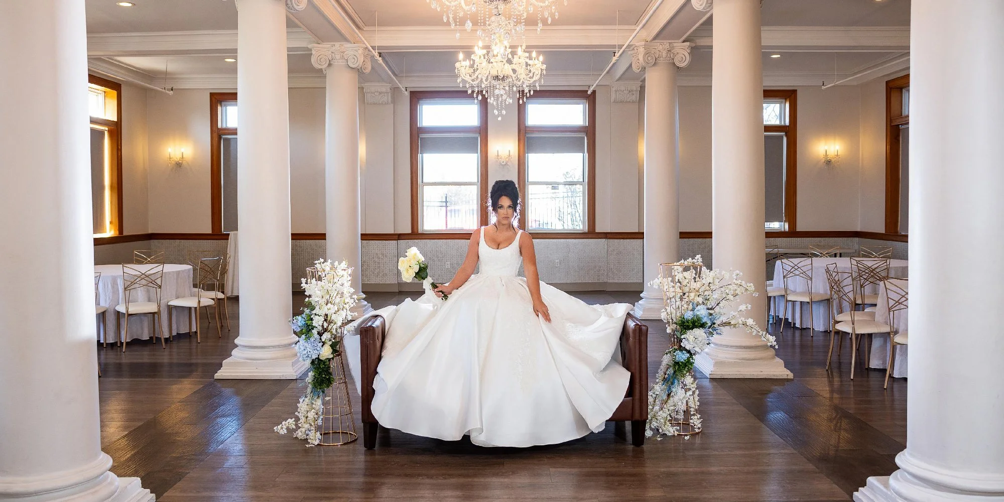 Bride in a white wedding gown holding a bouquet, seated on a couch in an elegant room with columns and chandeliers. Wedding Venue: The Loretto Events -- an upscale event venue and chapel located in Kansas City, Missouri