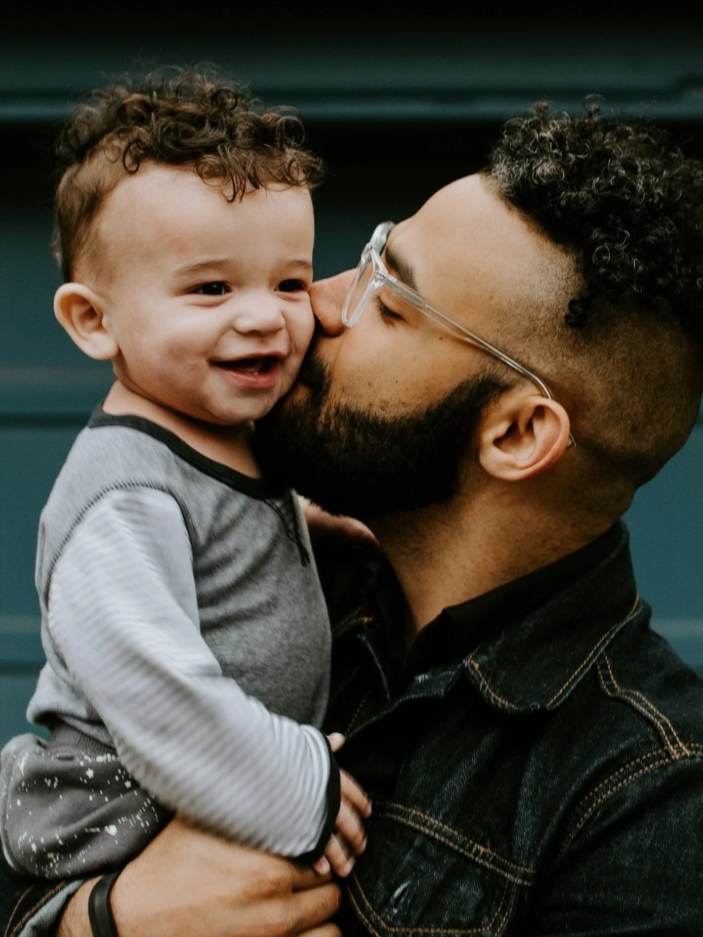 A man with glasses and a beard is holding a smiling young boy with curly hair, and the boy is giving the man a kiss on the nose.