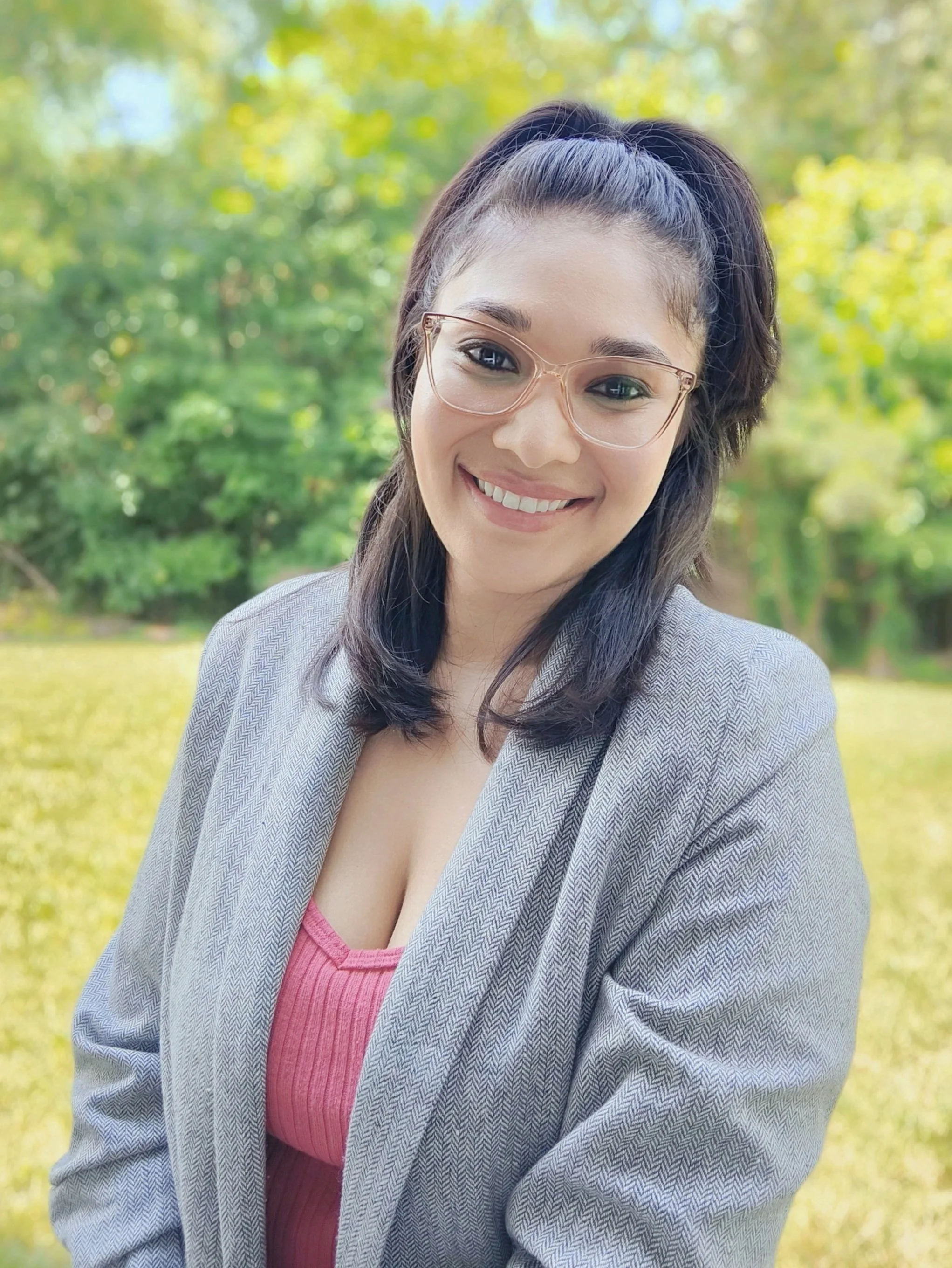 A young woman with glasses, wearing a grey blazer and pink top, smiling outdoors with green trees in the background.
