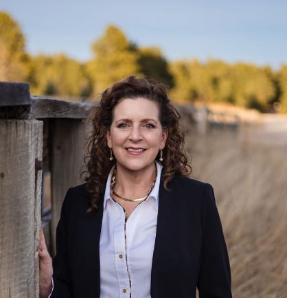 A woman with curly brown hair wearing a dark blazer and white shirt, standing outdoors near a wooden railing with trees and sky in the background.