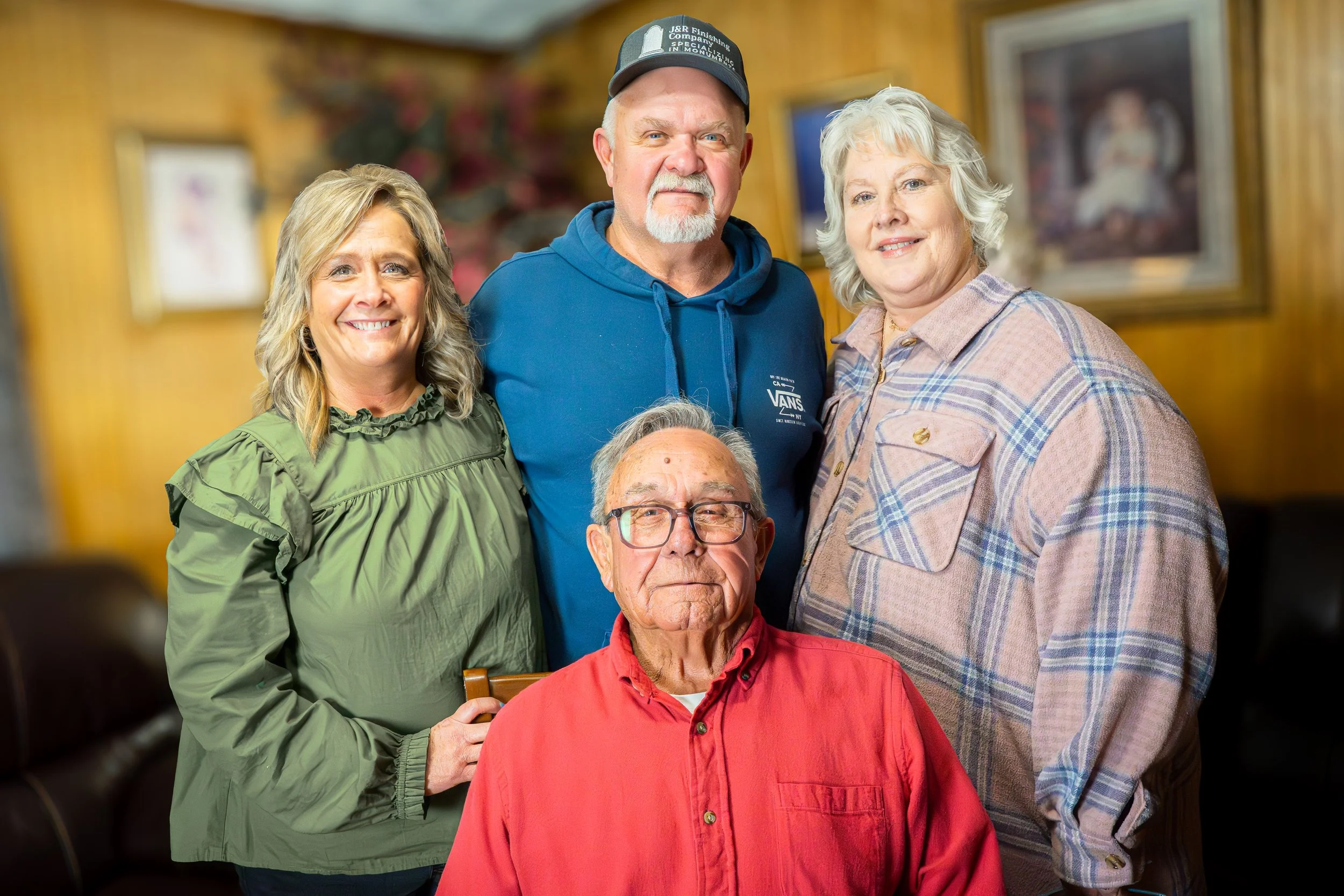 Group photo of three adults standing behind an elderly man seated indoors.