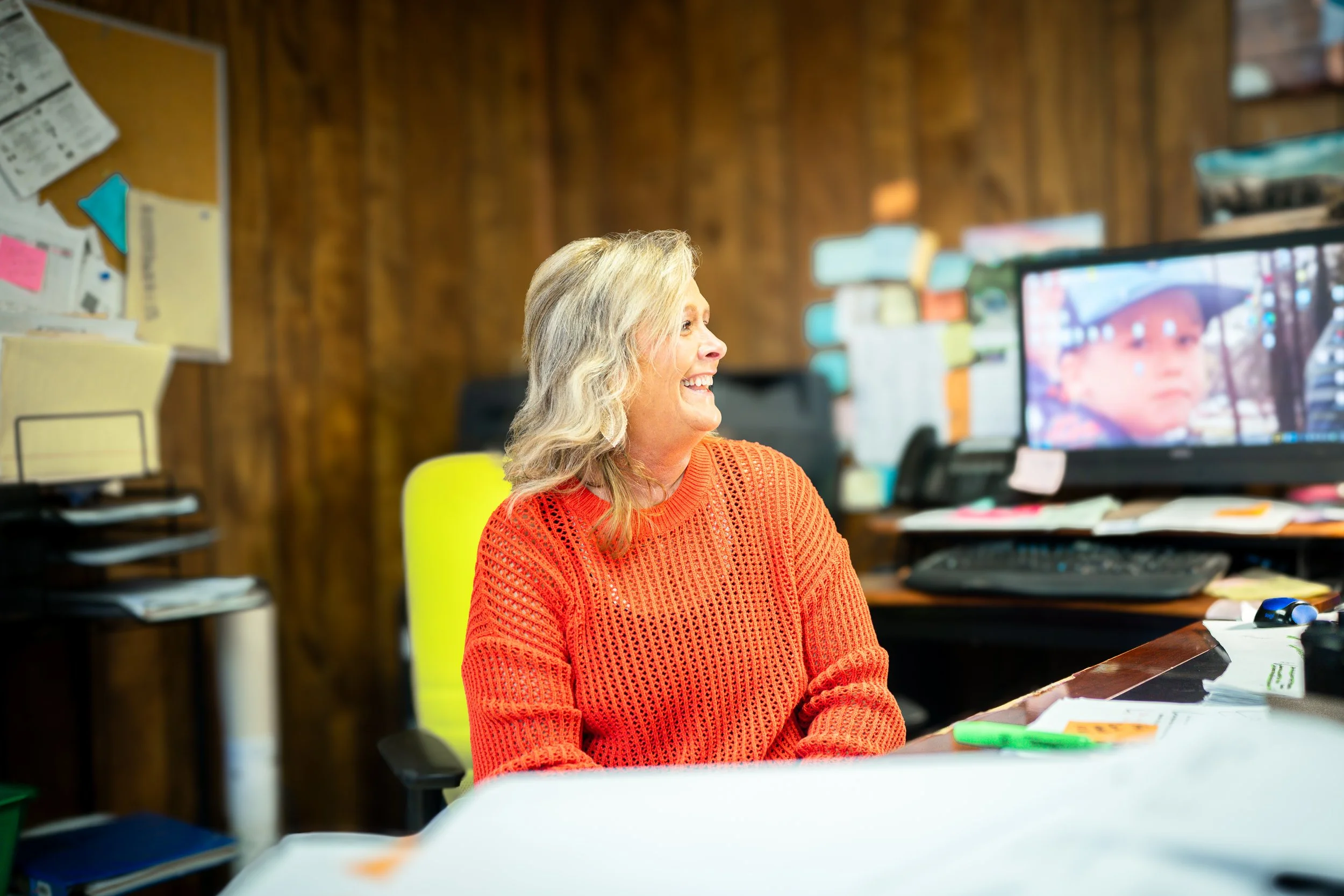 Woman in orange sweater smiling in an office with a cluttered desk and computer.