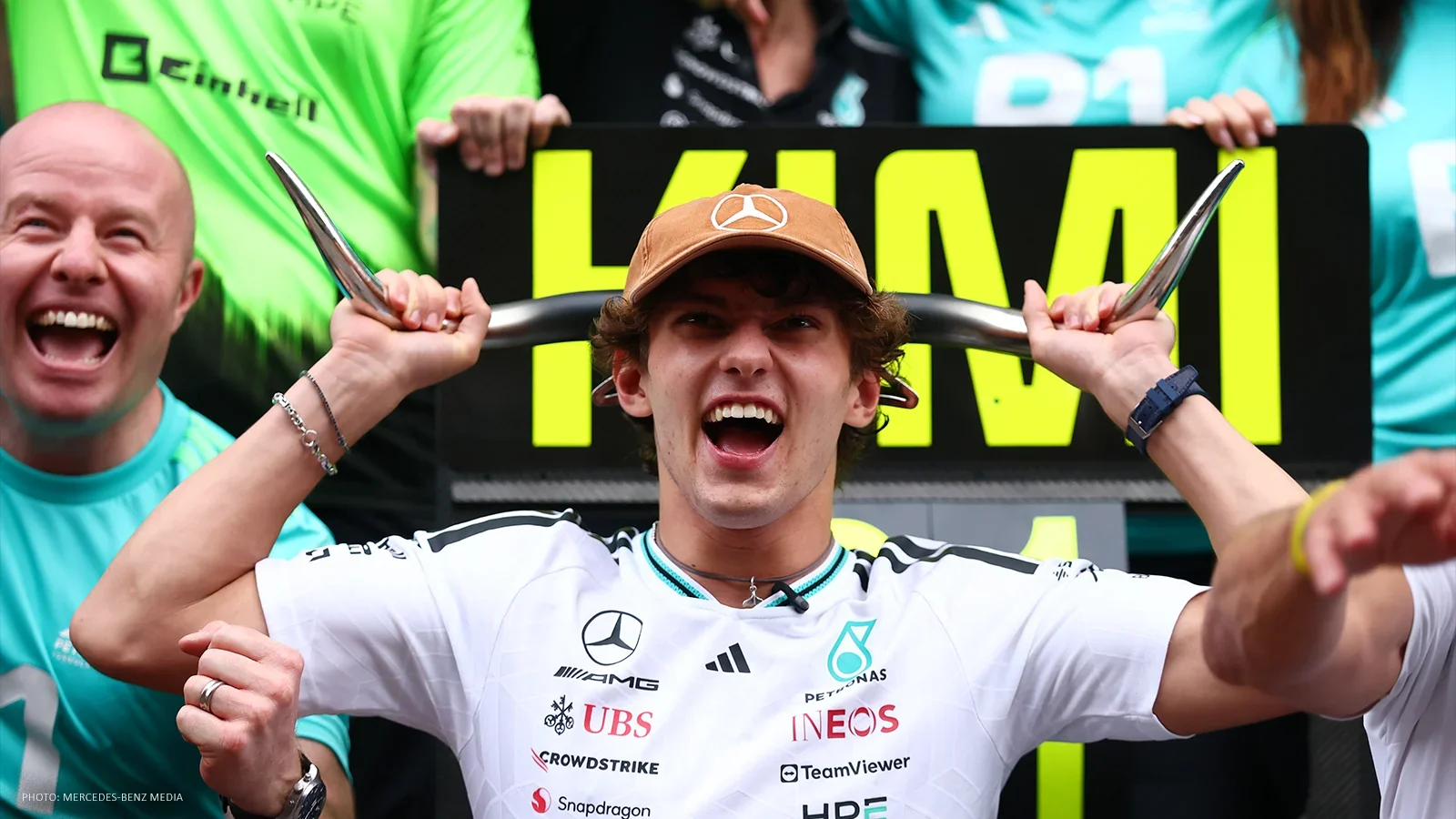 Kimi Antonelli smiling with metallic horns held behind his head, surrounded by Mercedes team members.