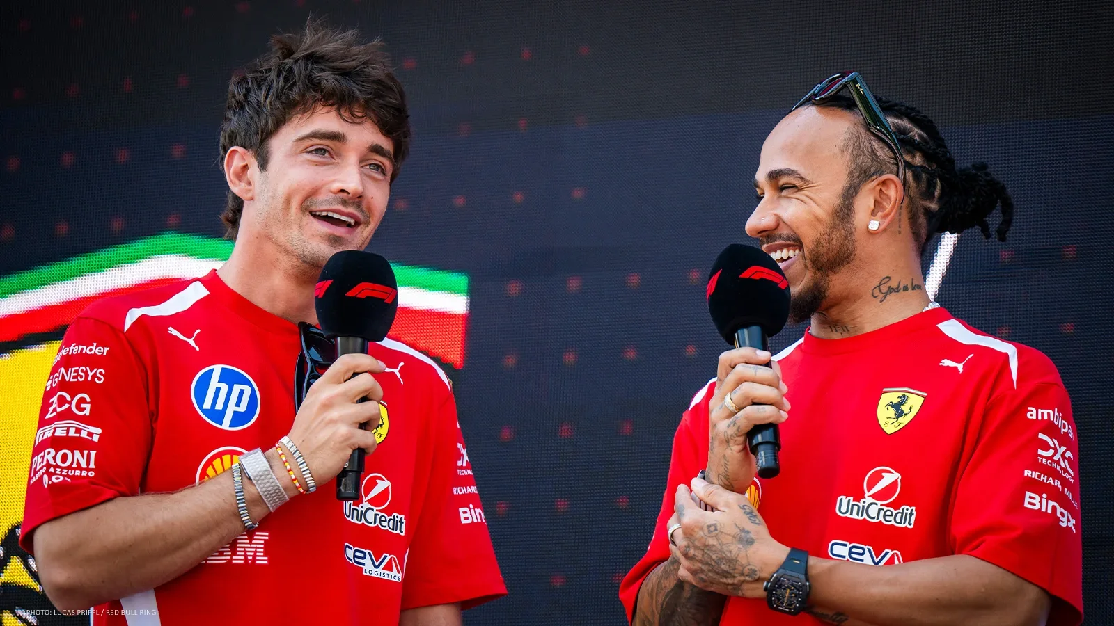 Charles Leclerc and Lewis Hamilton smiling while holding microphones during a Ferrari stage appearance