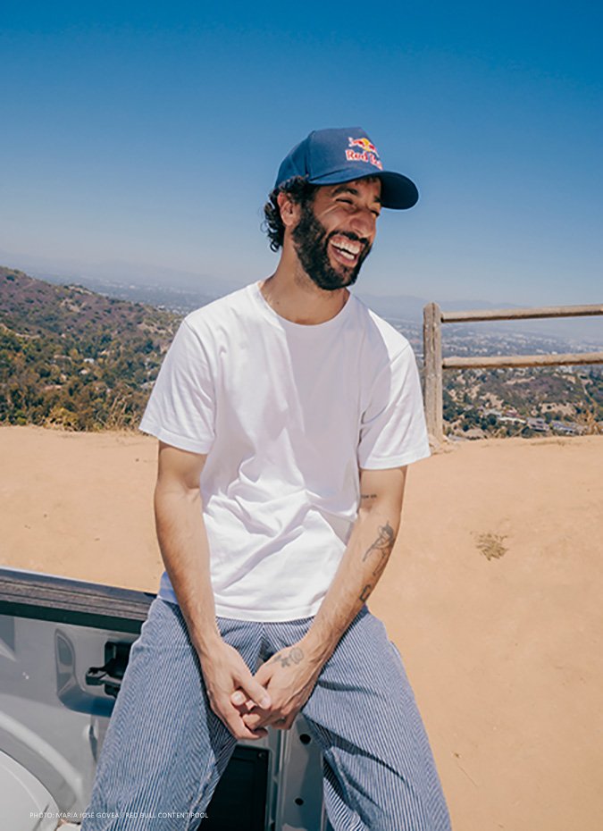 Daniel Ricciardo smiling outdoors in Los Angeles, wearing a white shirt and Red Bull cap.