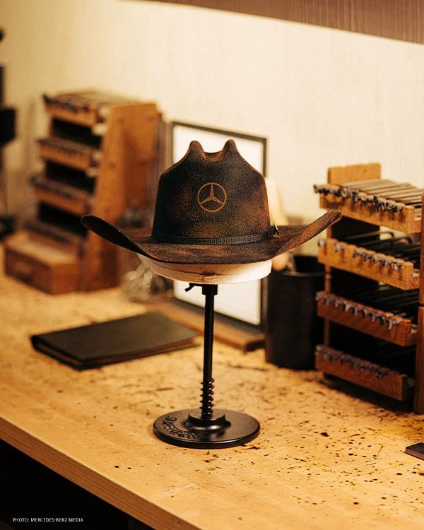 Black cowboy hat with an embroidered Mercedes star displayed on a stand.
