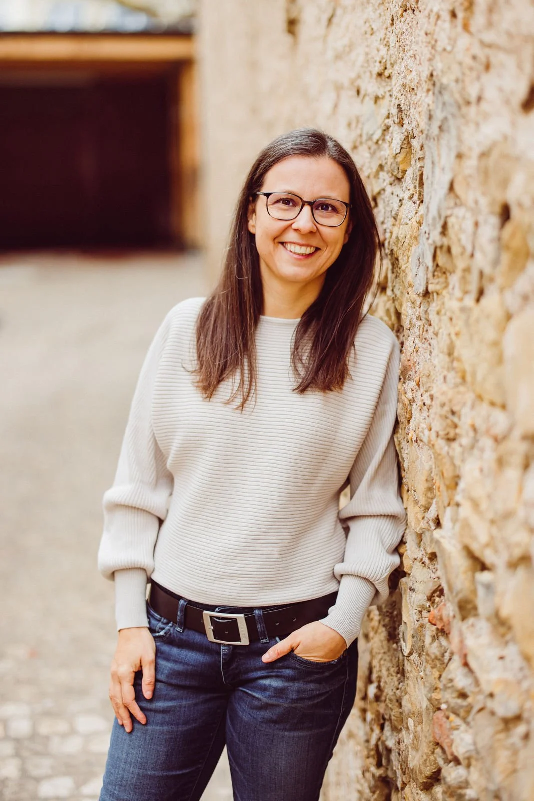 Tanja Heirens Coaching | Eine lächelnde Frau mit Brille lehnt an einer Steinmauer und trägt einen cremefarbenen Pullover sowie eine Jeans.