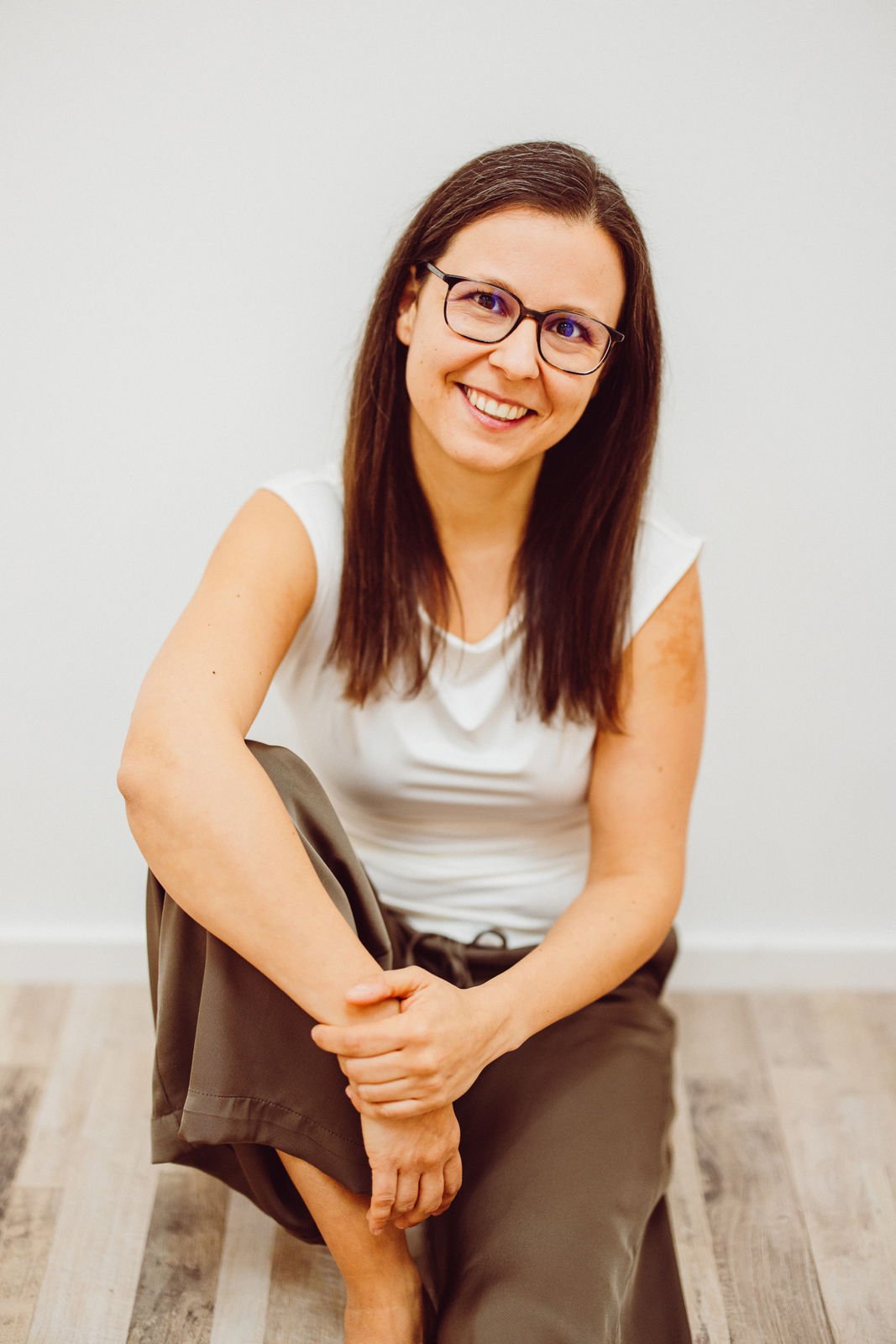Tanja Heirens Coaching | Frau mit Brille und langen Haaren, lächelnd, sitzt auf Holzboden, trägt ein weißes Oberteil und dunkle Hose.