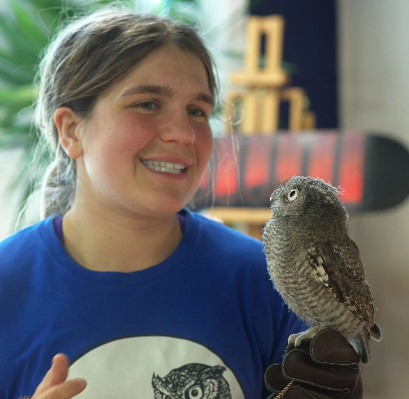 A young girl with light skin, brown hair, and blue eyes, smiling at the camera as she holds a small owl on her gloved hand indoors. The background is blurred with some green foliage and art supplies.