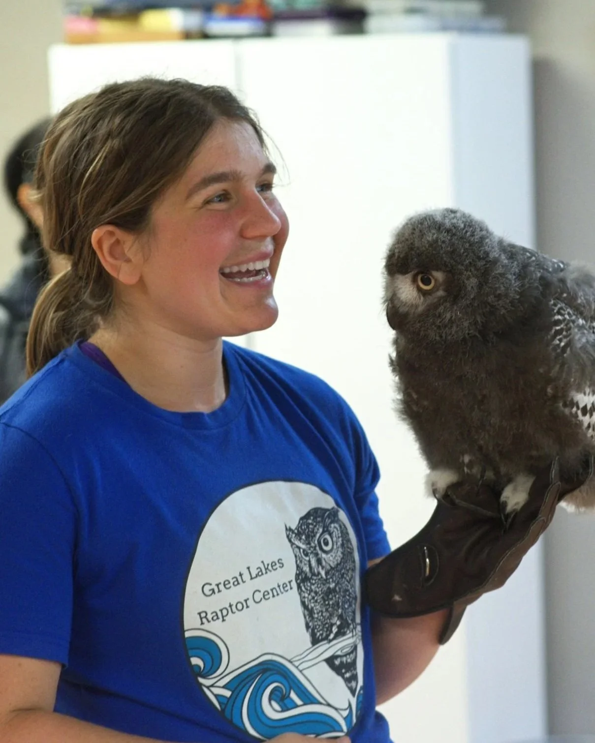 A woman holding an owl on her gloved hand, smiling and looking at the owl. She wears a blue t-shirt with a logo that says "Great Lakes Raptor Center" and features an owl illustration.