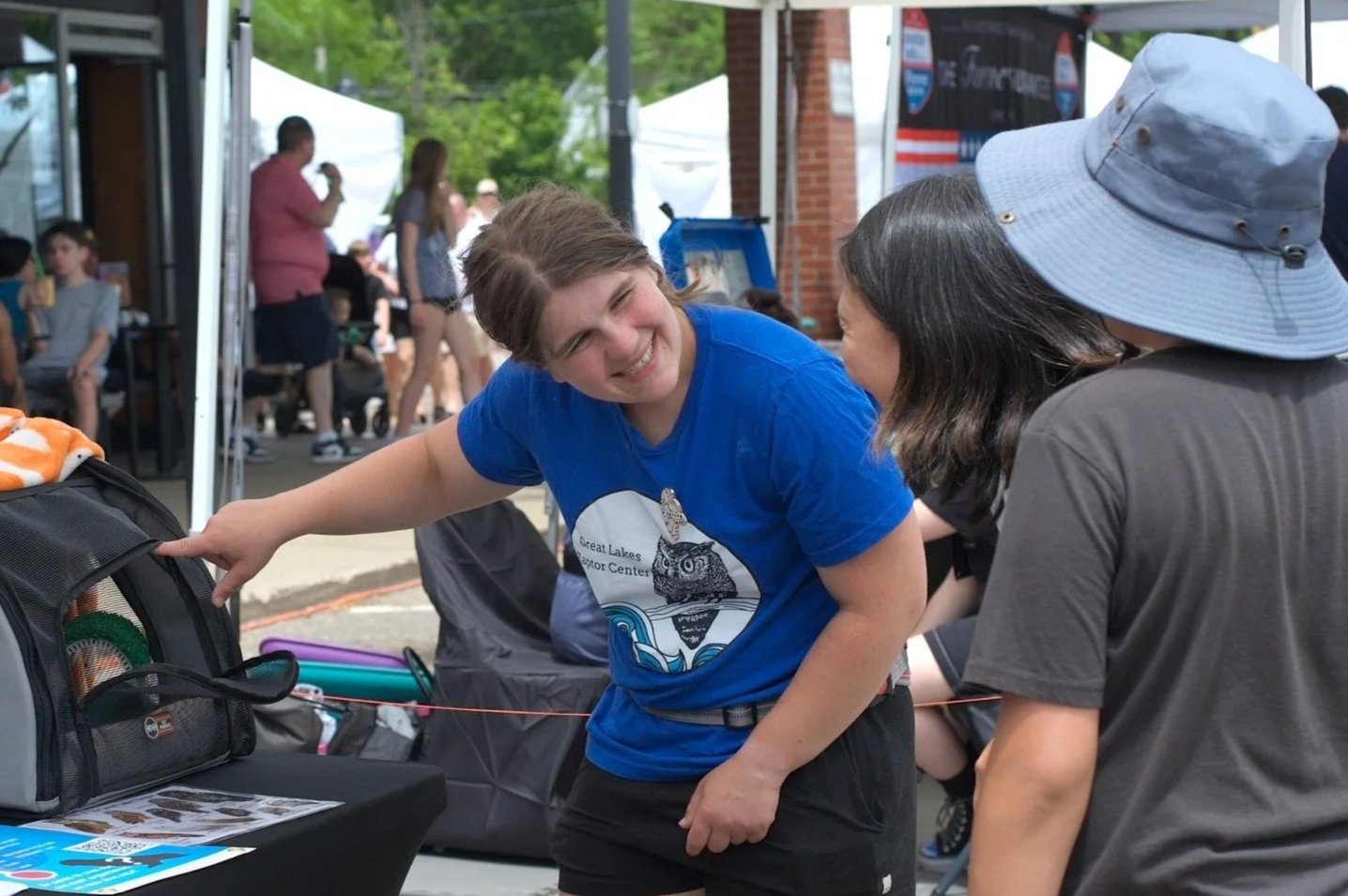People gathered outdoors at an event, with a young woman in a blue shirt smiling and talking to a woman wearing a sun hat. There are tents and other people in the background.