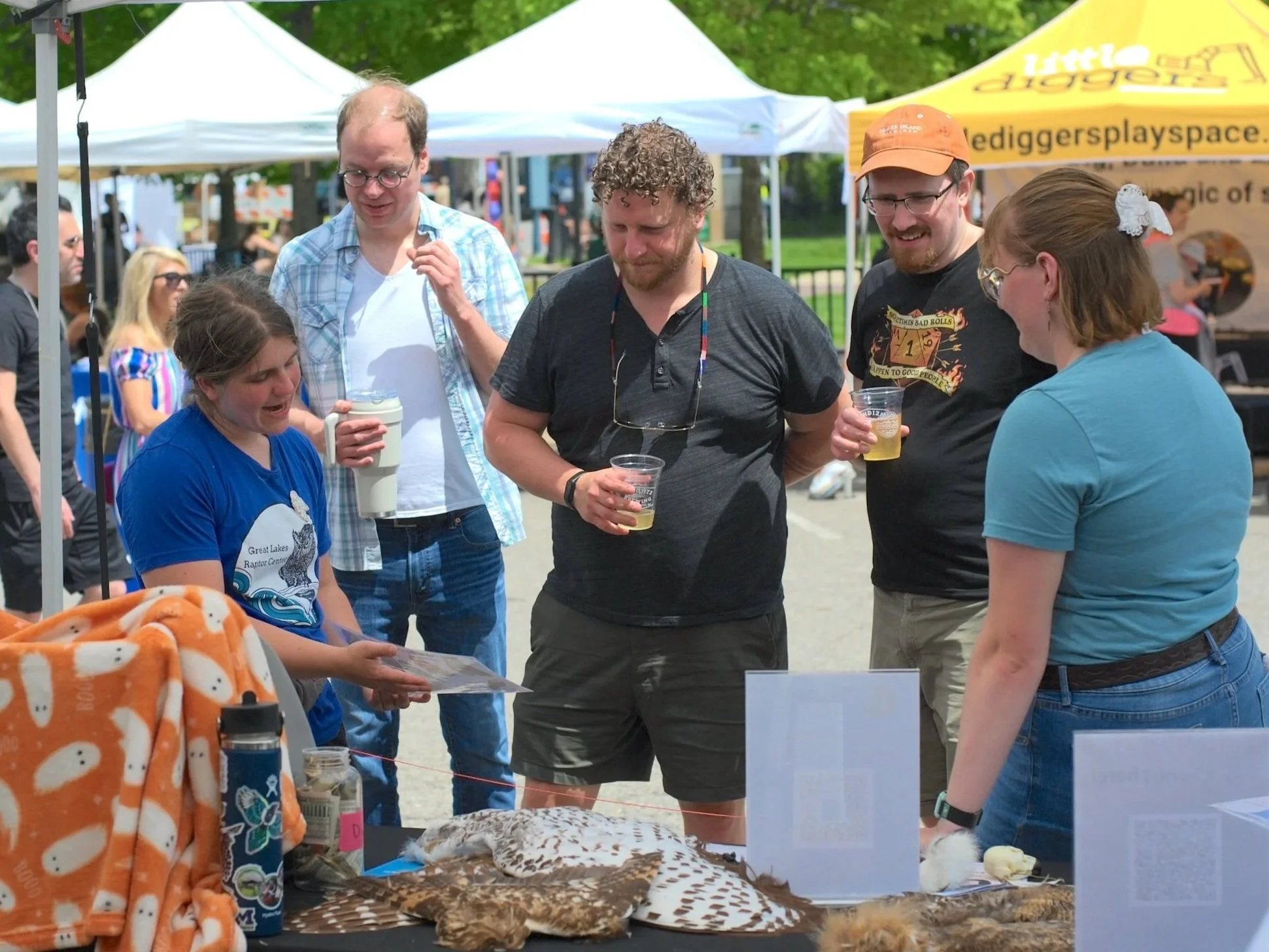 People are gathered around a booth at an outdoor event. Other tented booths are visible in the background. A woman in a blue shirt is giving information to three men and a woman, pointing to a laminated sheet of paper. Feathers lay on the table.