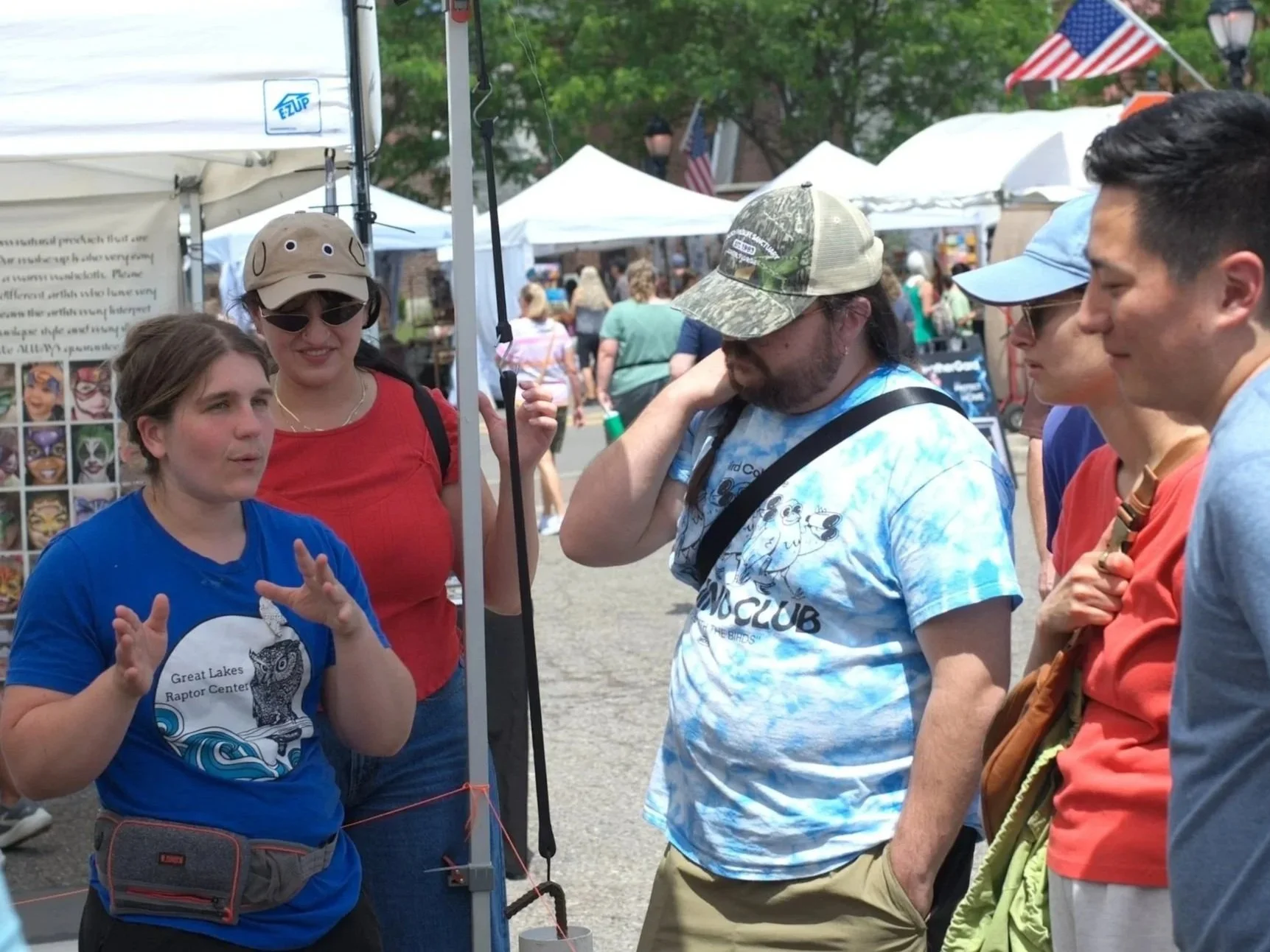 Group of people gathered outdoors at a festival or fair, engaging in conversation. Tents and pedestrians are visible in the background.