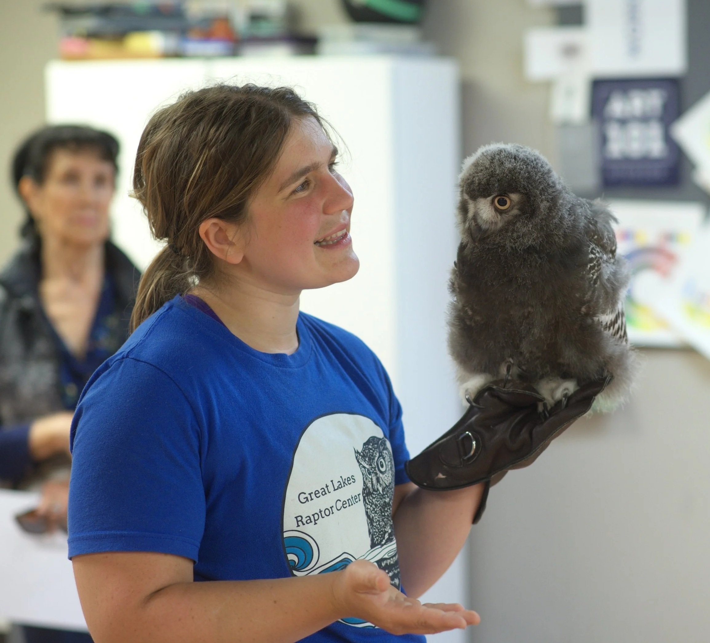 A woman holding a young owl on her gloved hand at an indoor event, with a blurred background of other people and equipment.