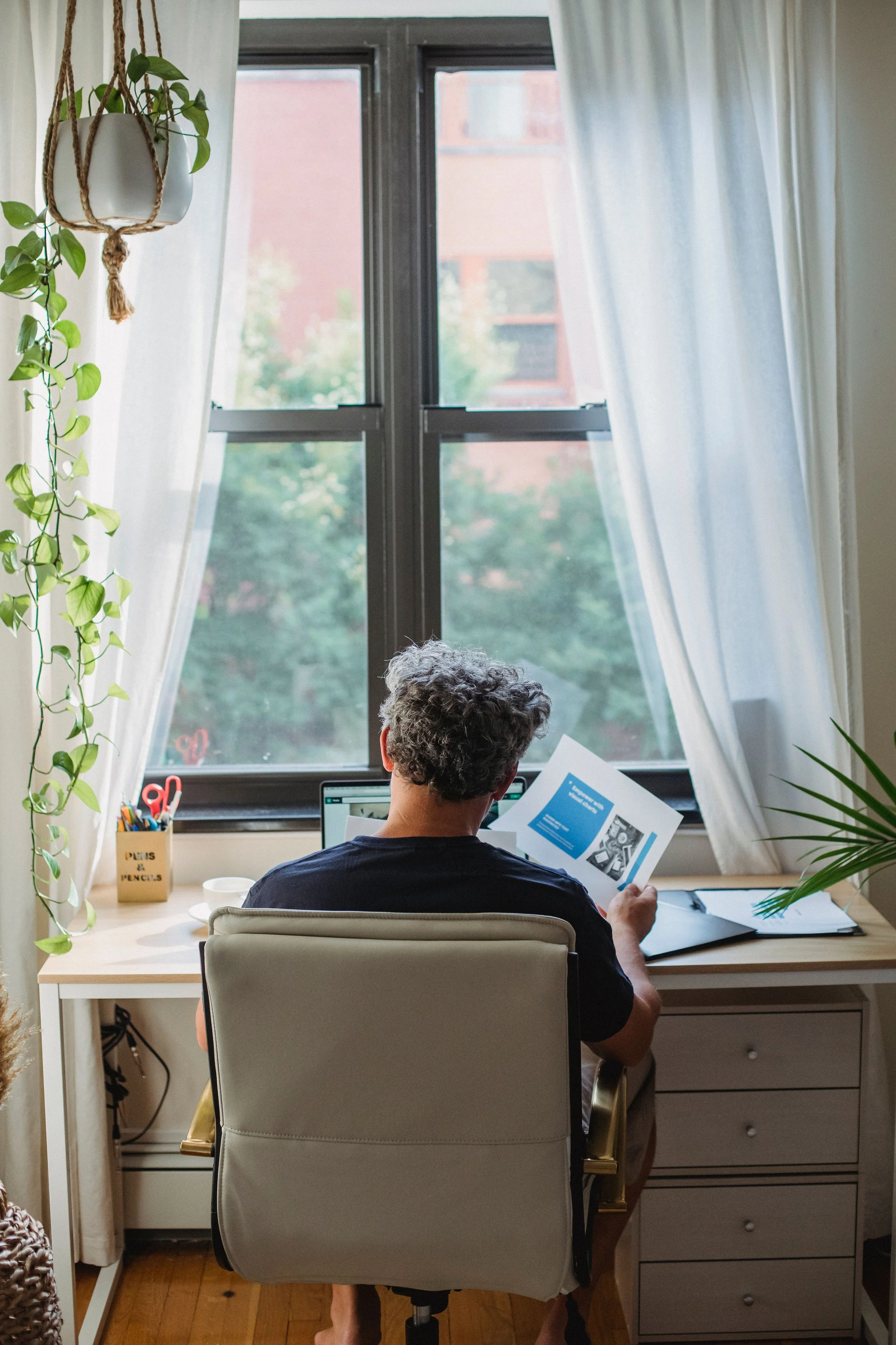 Person working at a desk by a window, holding printed papers, surrounded by plants and sheer curtains at Yarrow Functional Wellness with Amy Stetson