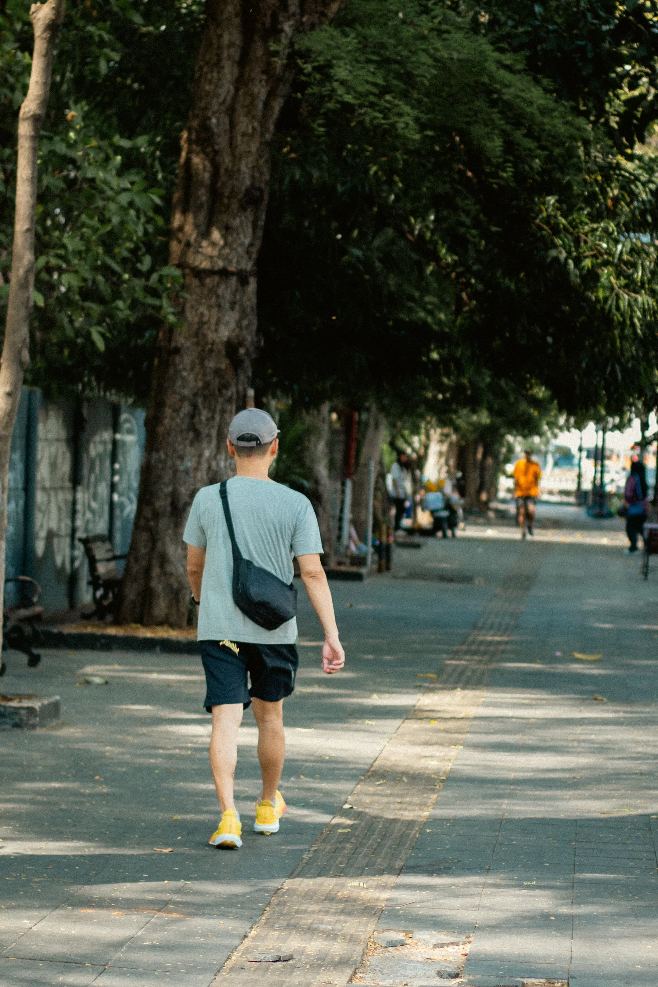 Person in a gray cap walking along a tree-lined urban sidewalk with a shoulder bag at Yarrow Functional Wellness with Amy Stetson