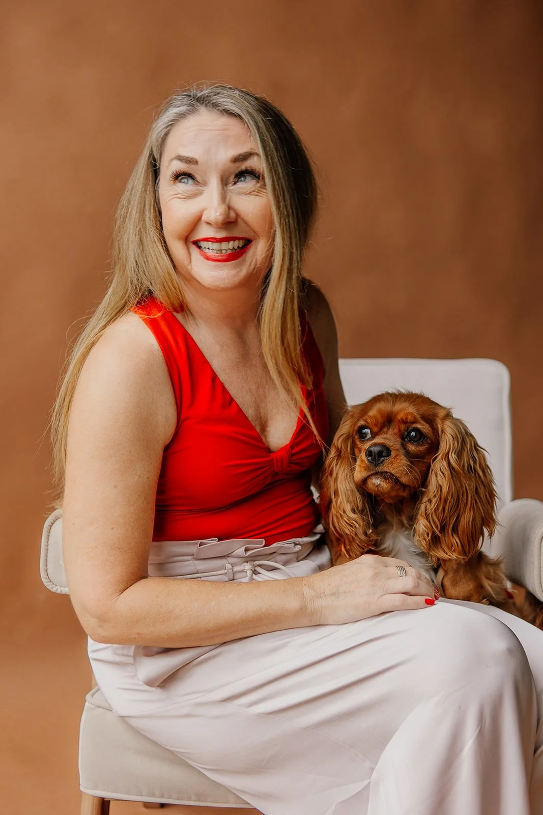A woman in a red top and white skirt sits on a chair, smiling at a small dog beside her.