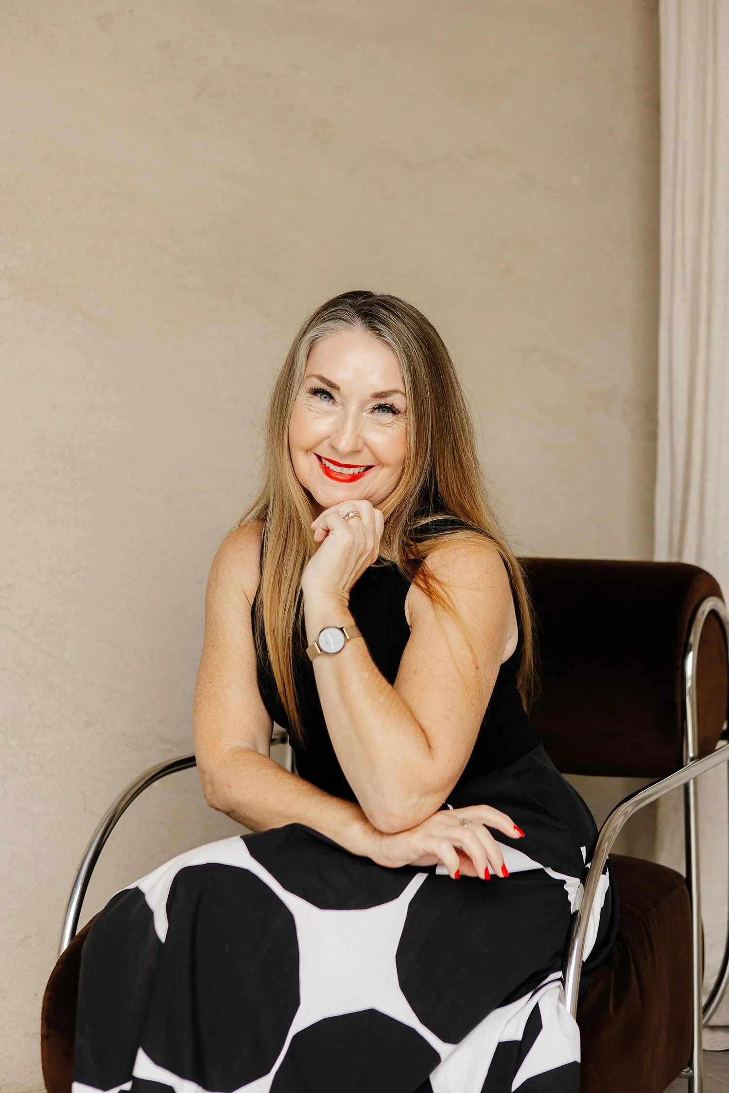Smiling woman with long hair sits in a chair, wearing a black top and polka dot skirt, with her hand resting on her chin.