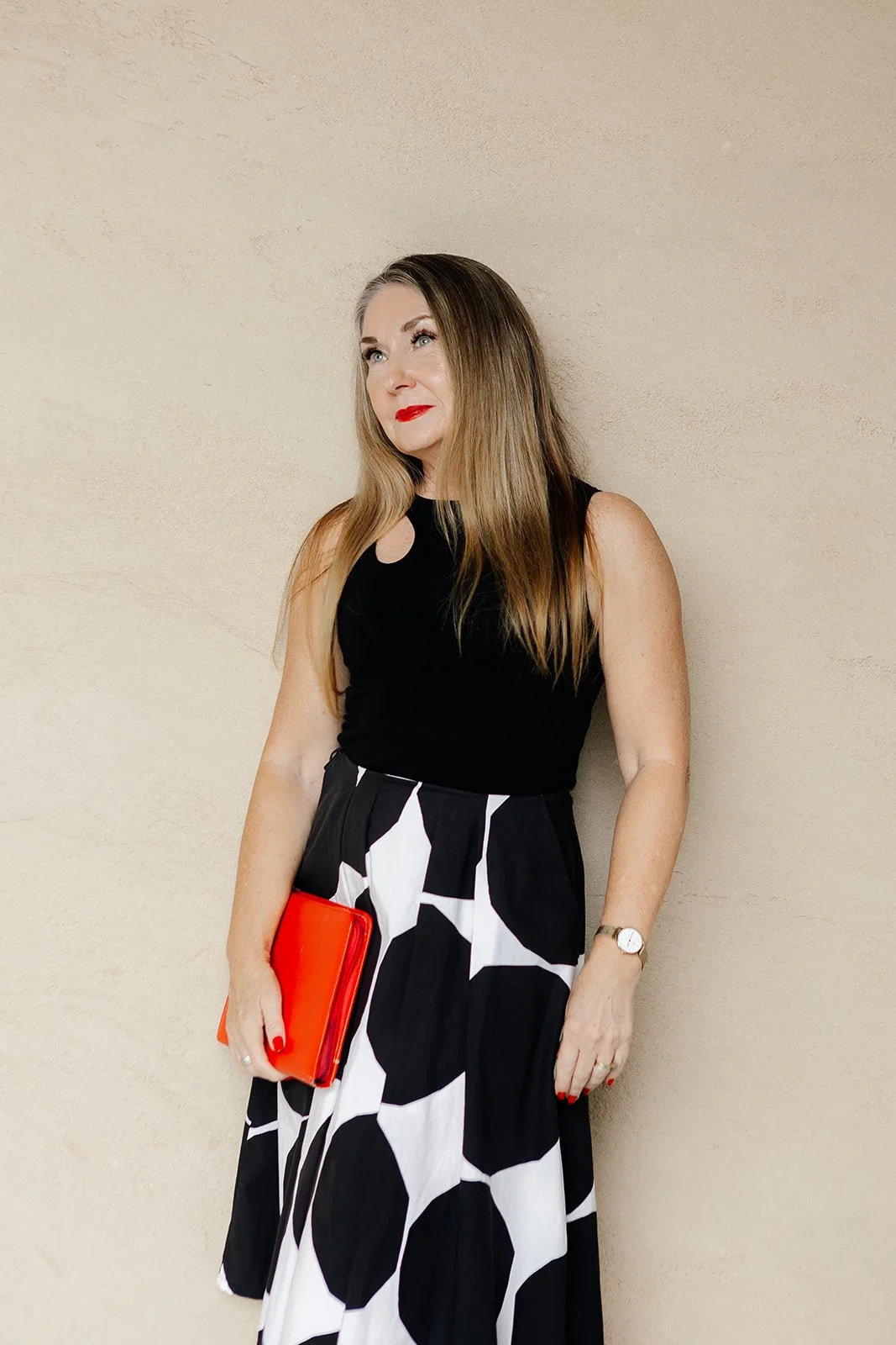 A woman wearing a black and white polka dot dress holds a vibrant red clutch, smiling confidently at the camera.