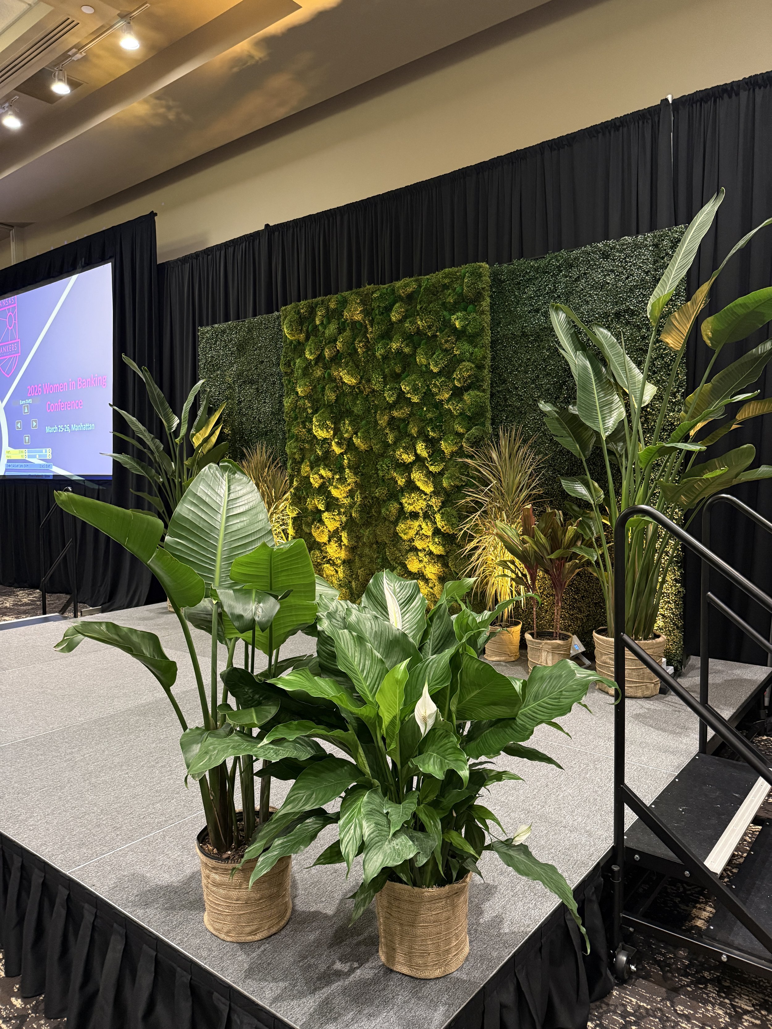 Stage decorated with green plants and a moss wall at an indoor event, with a large screen displaying information about the 2026 Wellness coalition of Wichita conference.