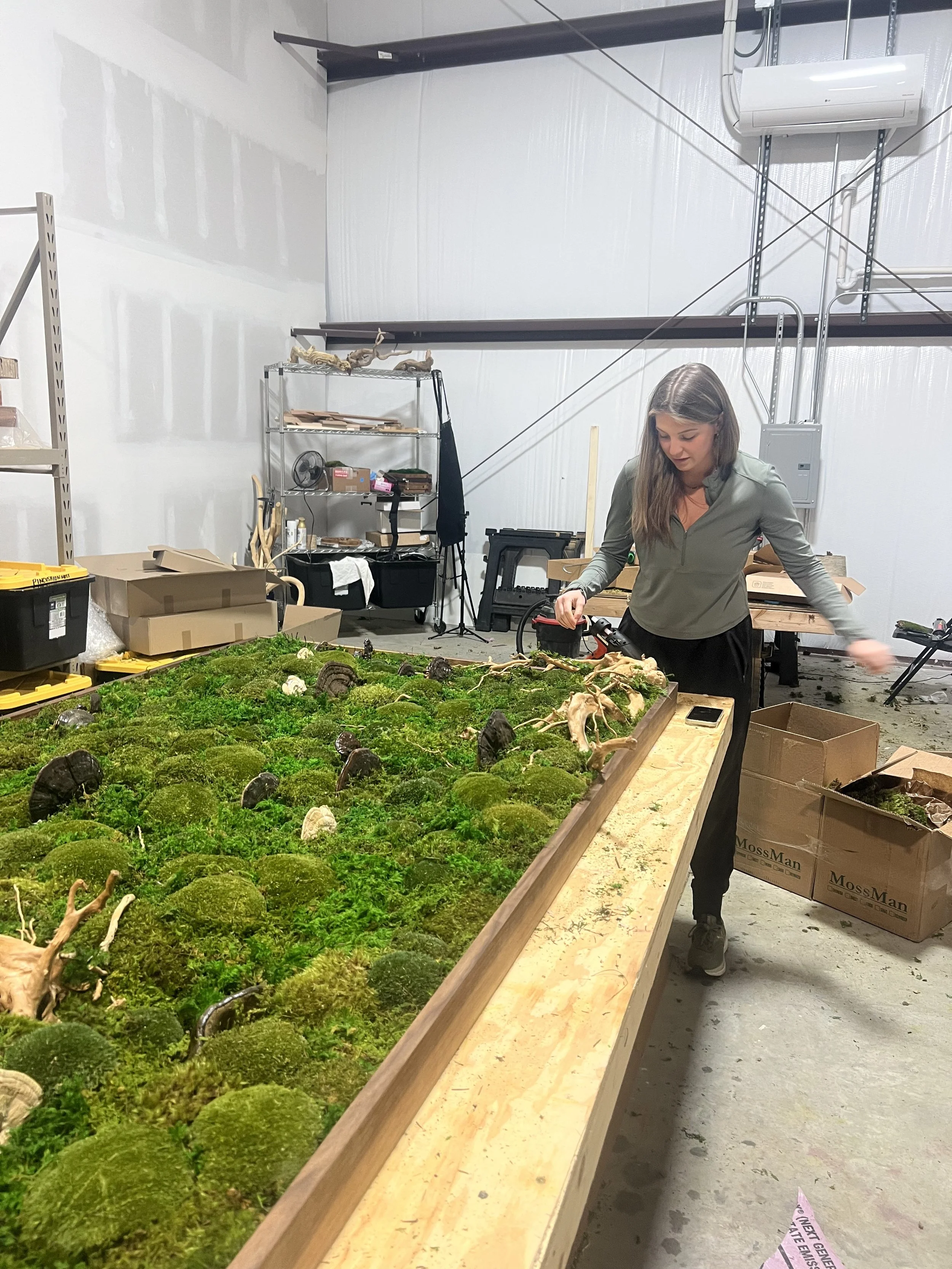 A woman working on a moss and skeleton art project in a workshop with shelves and boxes in the background.