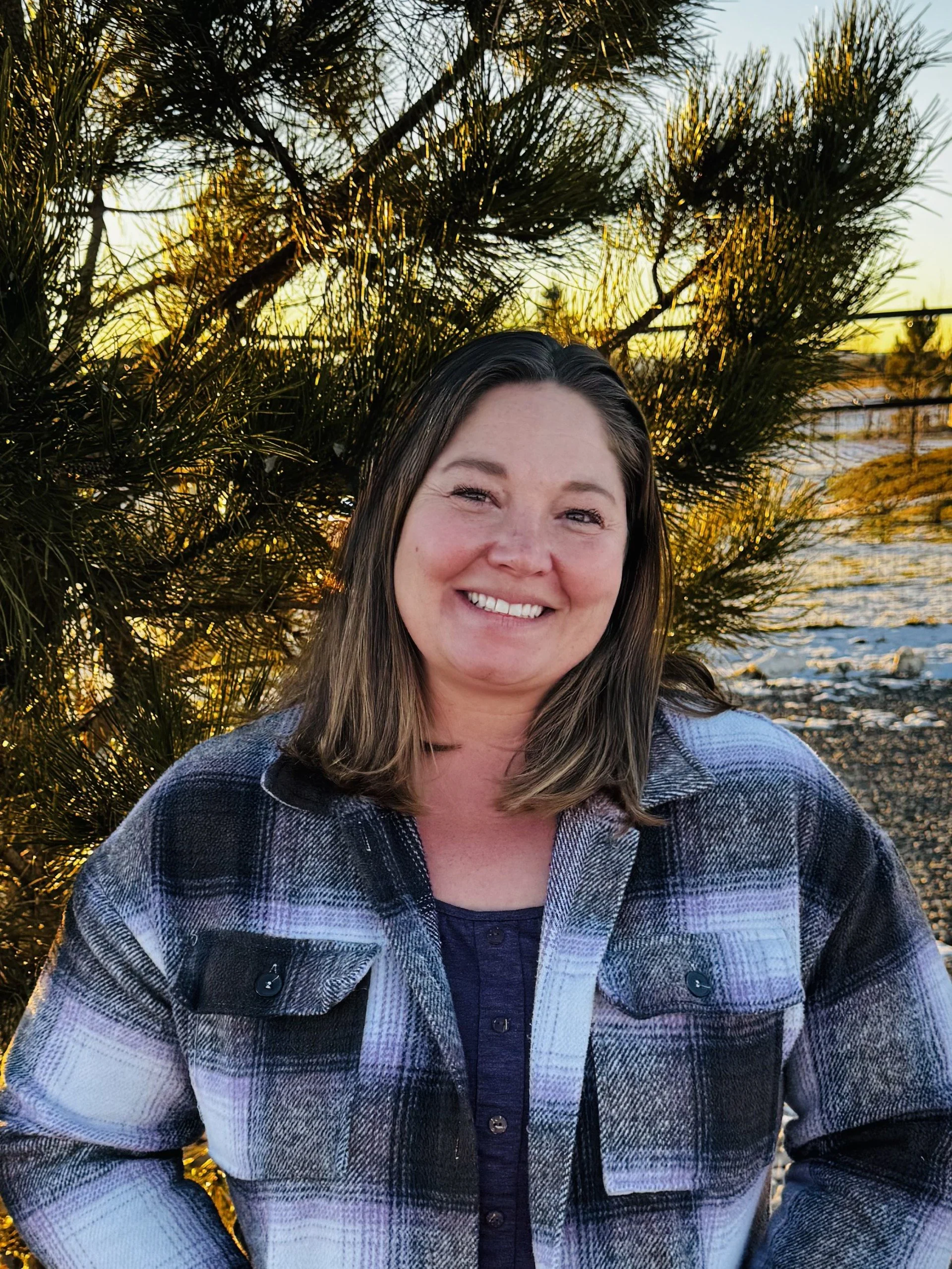 A woman smiling outdoors at sunset, standing in front of a pine tree and water.