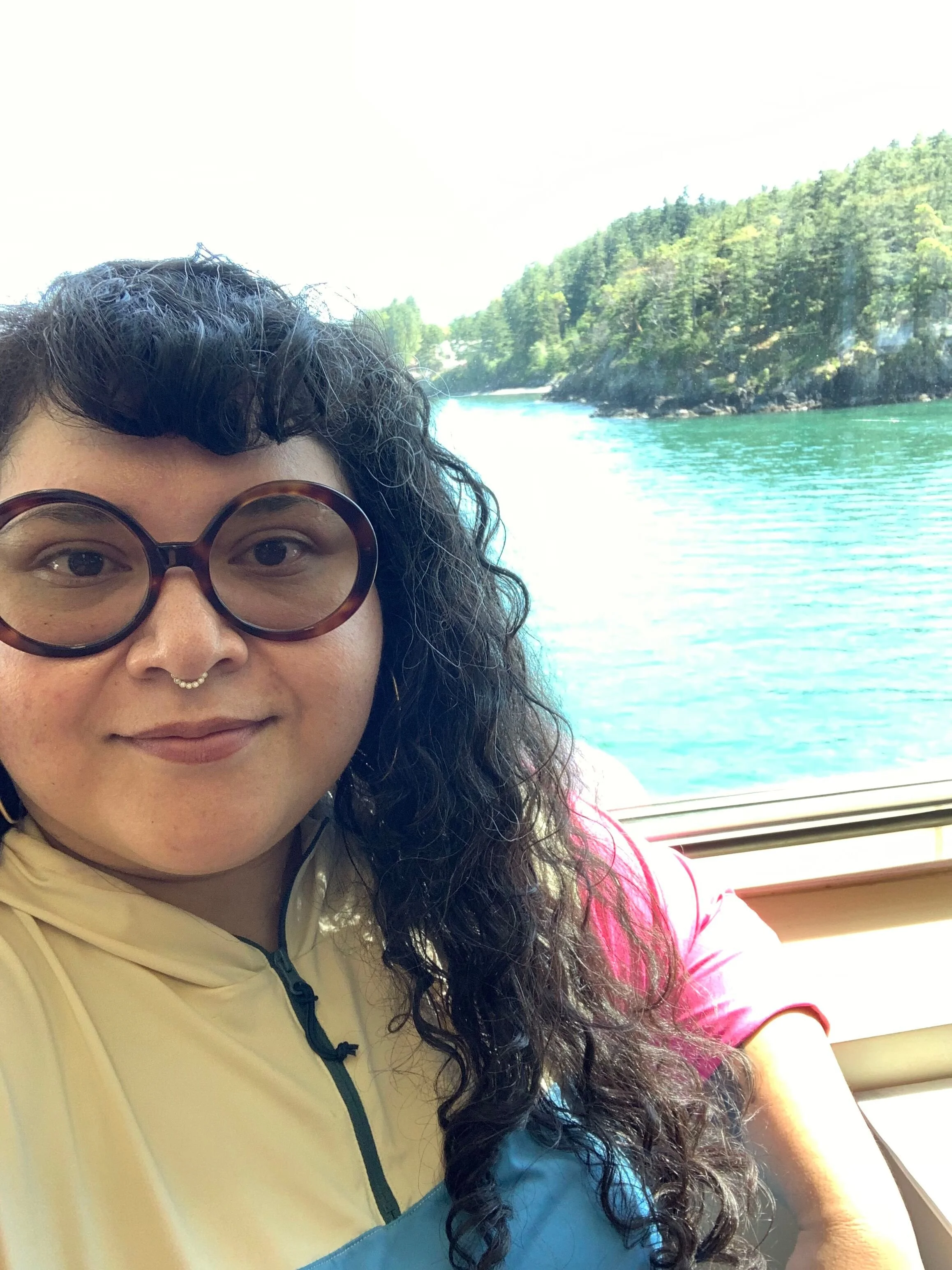 A person with curly hair, glasses, and a septum piercing taking a selfie on a boat with a river and forested hills in the background.